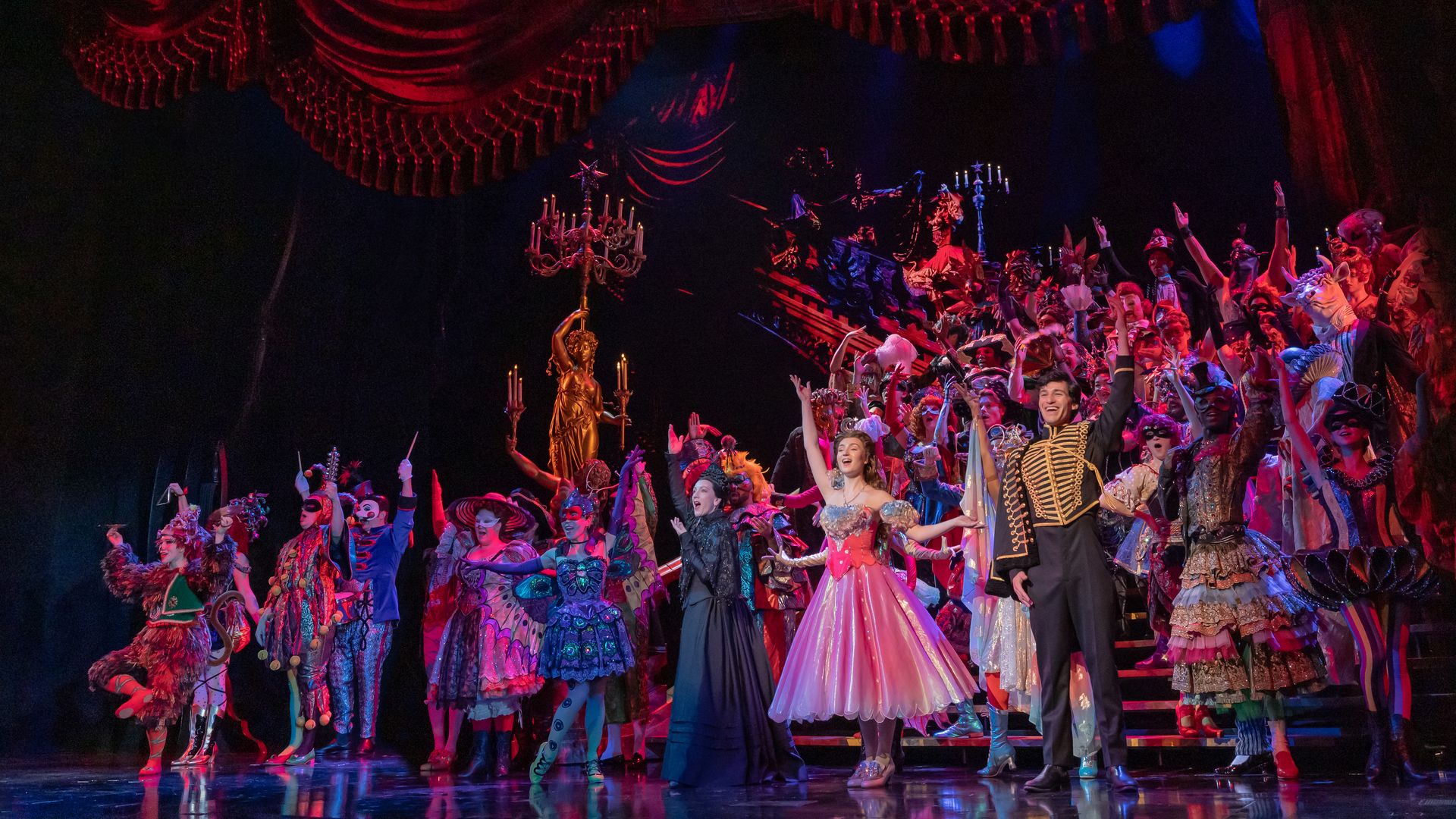 Vibrant stage finale with a sea of performers in ornate costumes; a pink princess and a man in a gold-trimmed coat stand center as the chorus cheers on tiered steps under red velvet drapes.