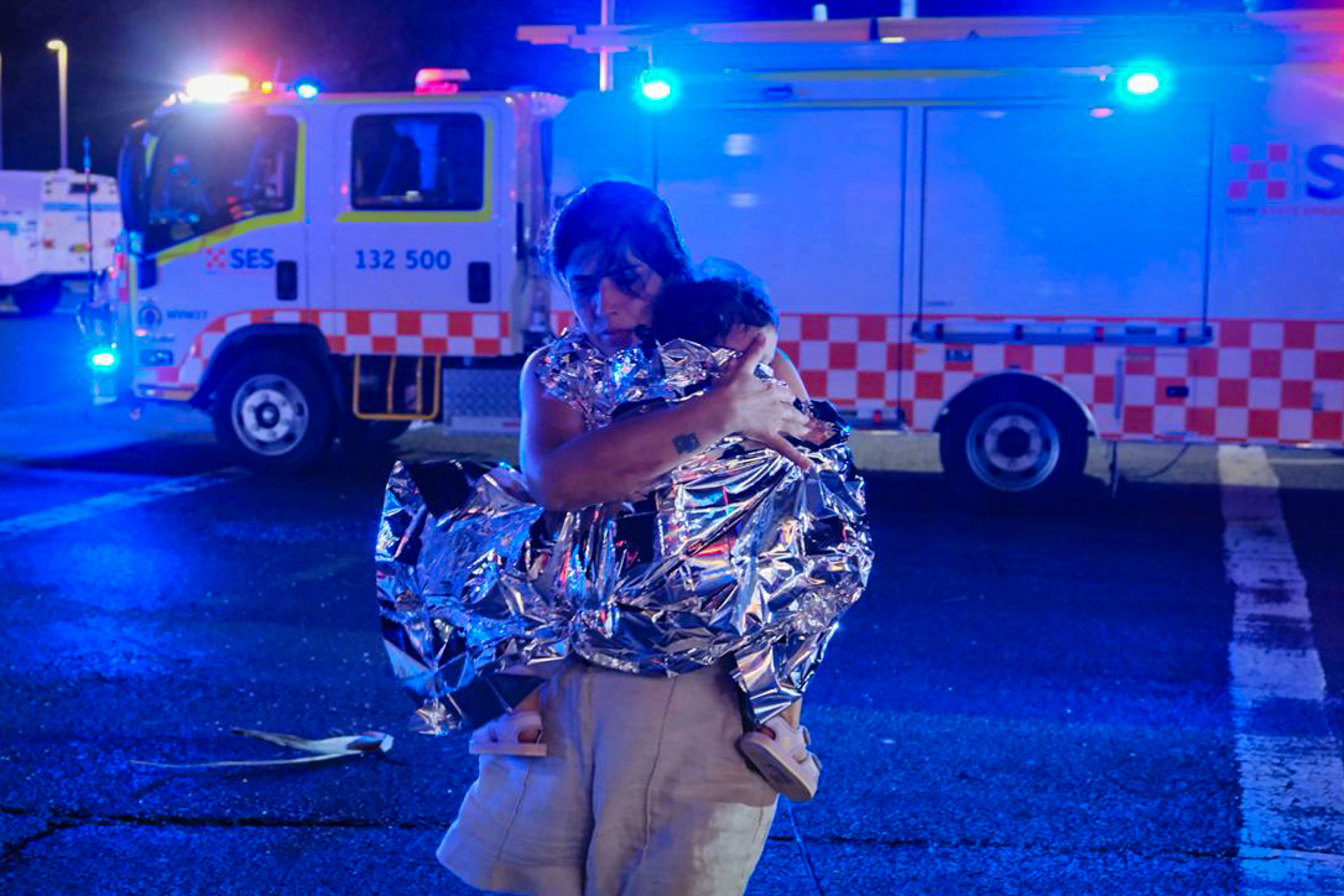 A member of the public leaves the scene with her child, who is covered in an emergency blanket, after a shooting at Bondi Beach on December 14, 2025 in Sydney, Australia. Two gunmen dressed in black fired several shots at Sydney's world-famous Bondi Beach, causing at least 10 injuries and three deat