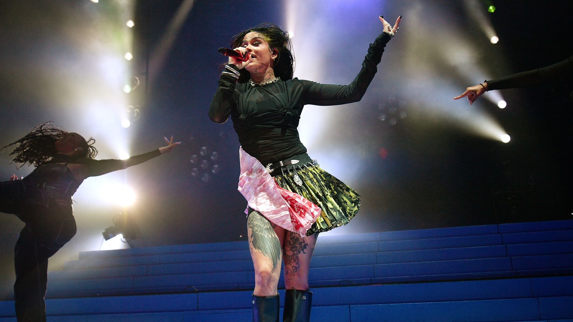 Kehlani performing in a pink and green skirt with a black top and stage lights behind her on a dark stage