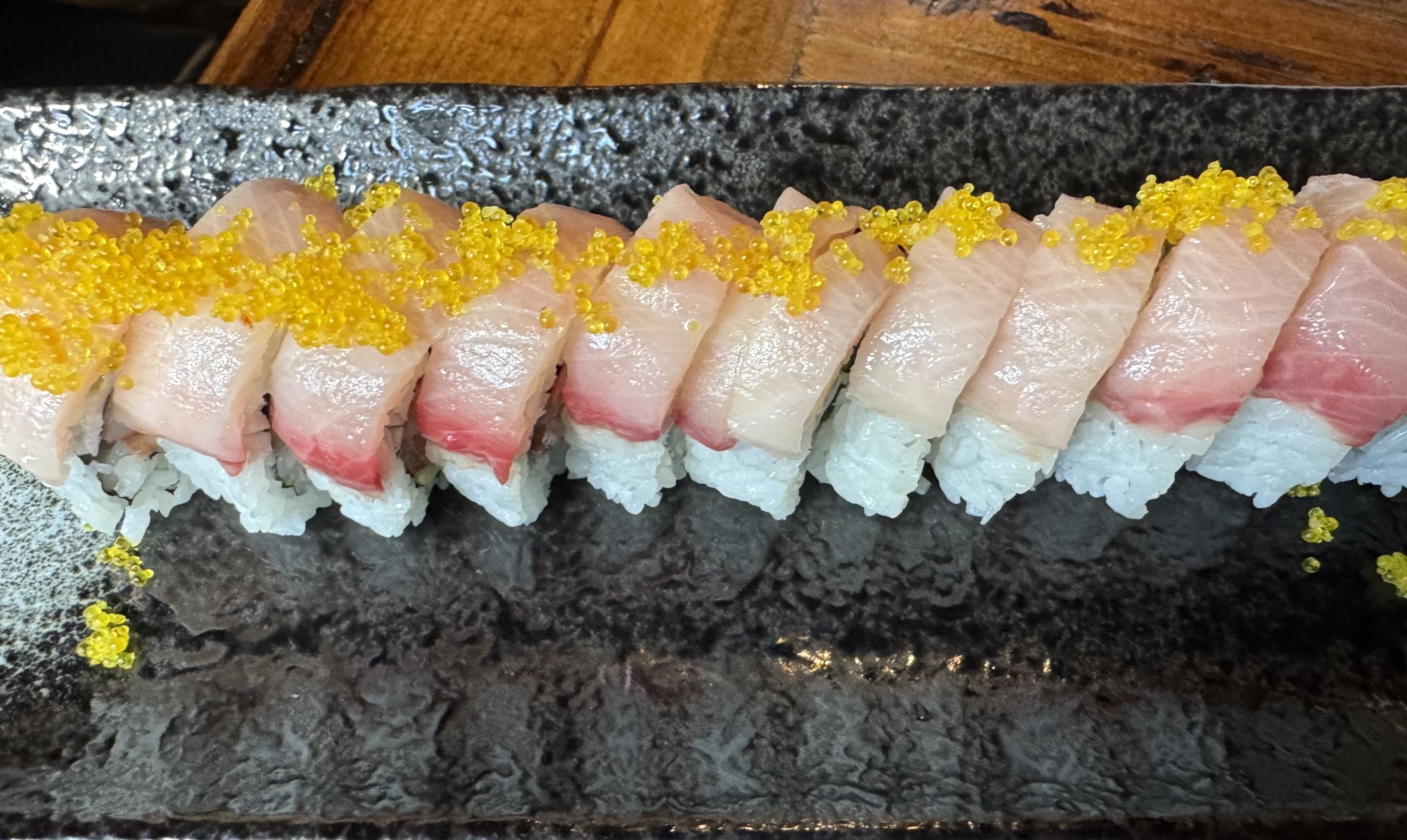 A sushi roll topped with slices of yellowtail and bright yellow tobiko (fish roe) is neatly arranged on a black ceramic plate.