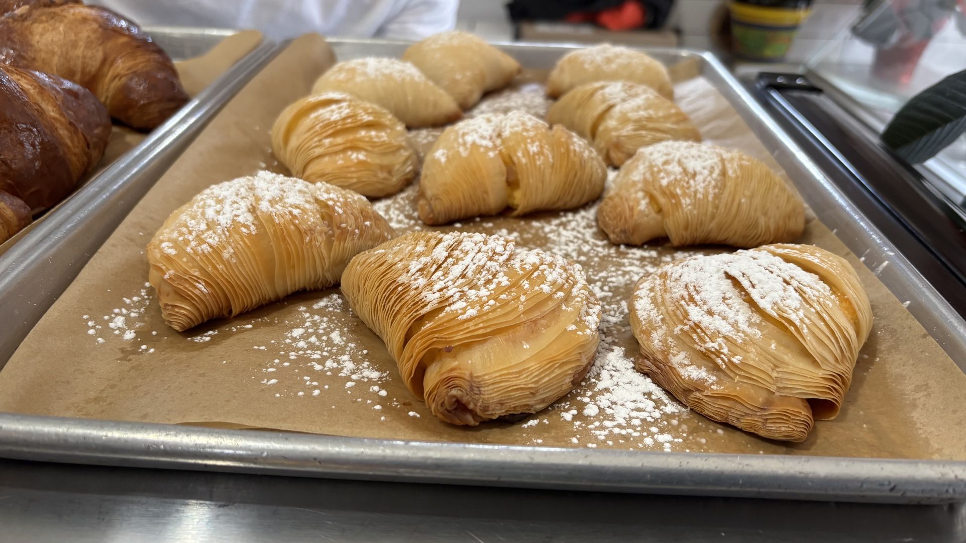 Tray of golden-brown flaky pastries dusted with powdered sugar on brown parchment paper, with a stainless steel tray of darker croissants partially visible on the left side.