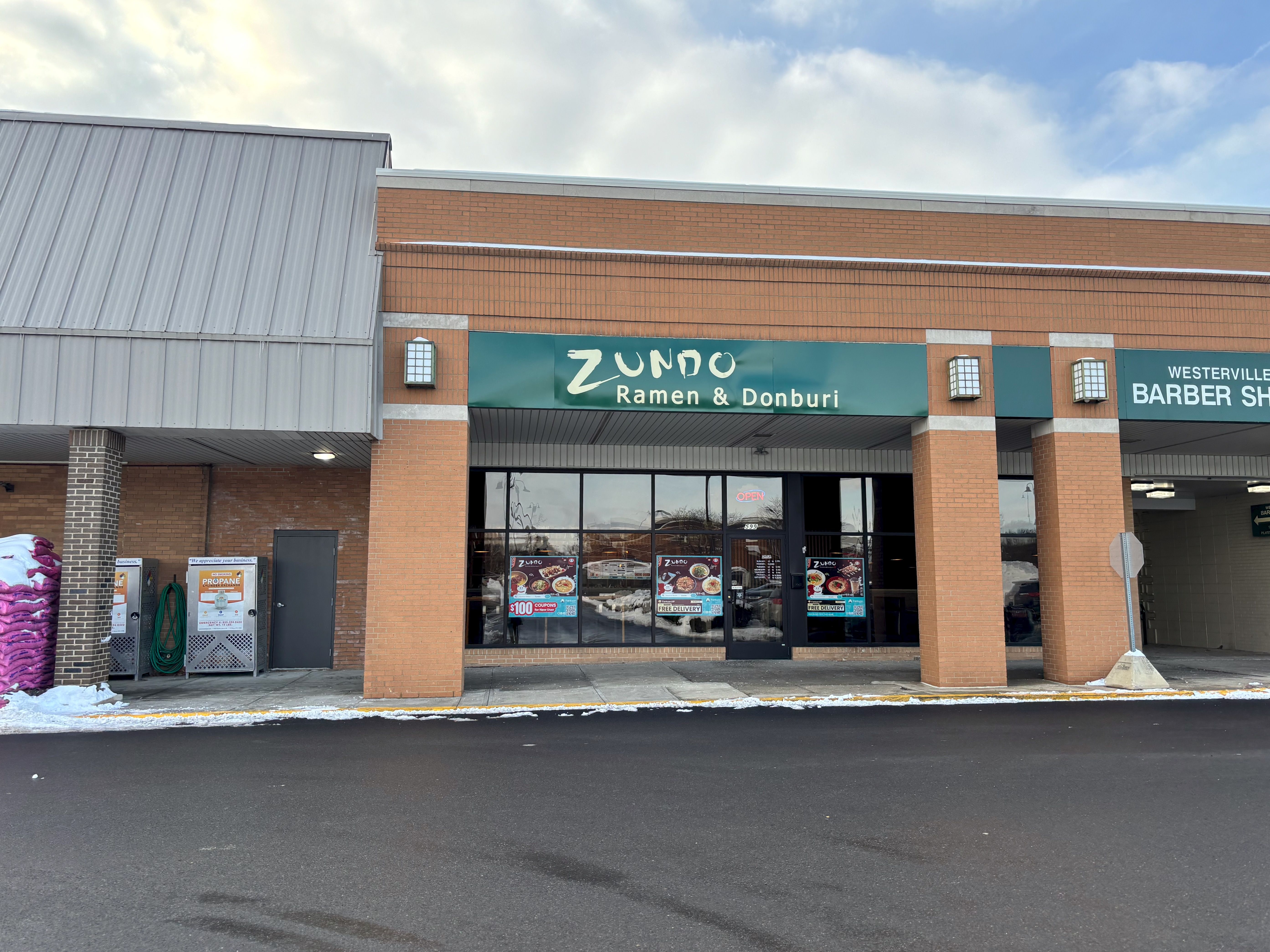 Exterior of a brick shopping plaza with Zundo Ramen & Donburi restaurant in the center, green sign, snow on sidewalks, and a barber shop to the right under cloudy sky.