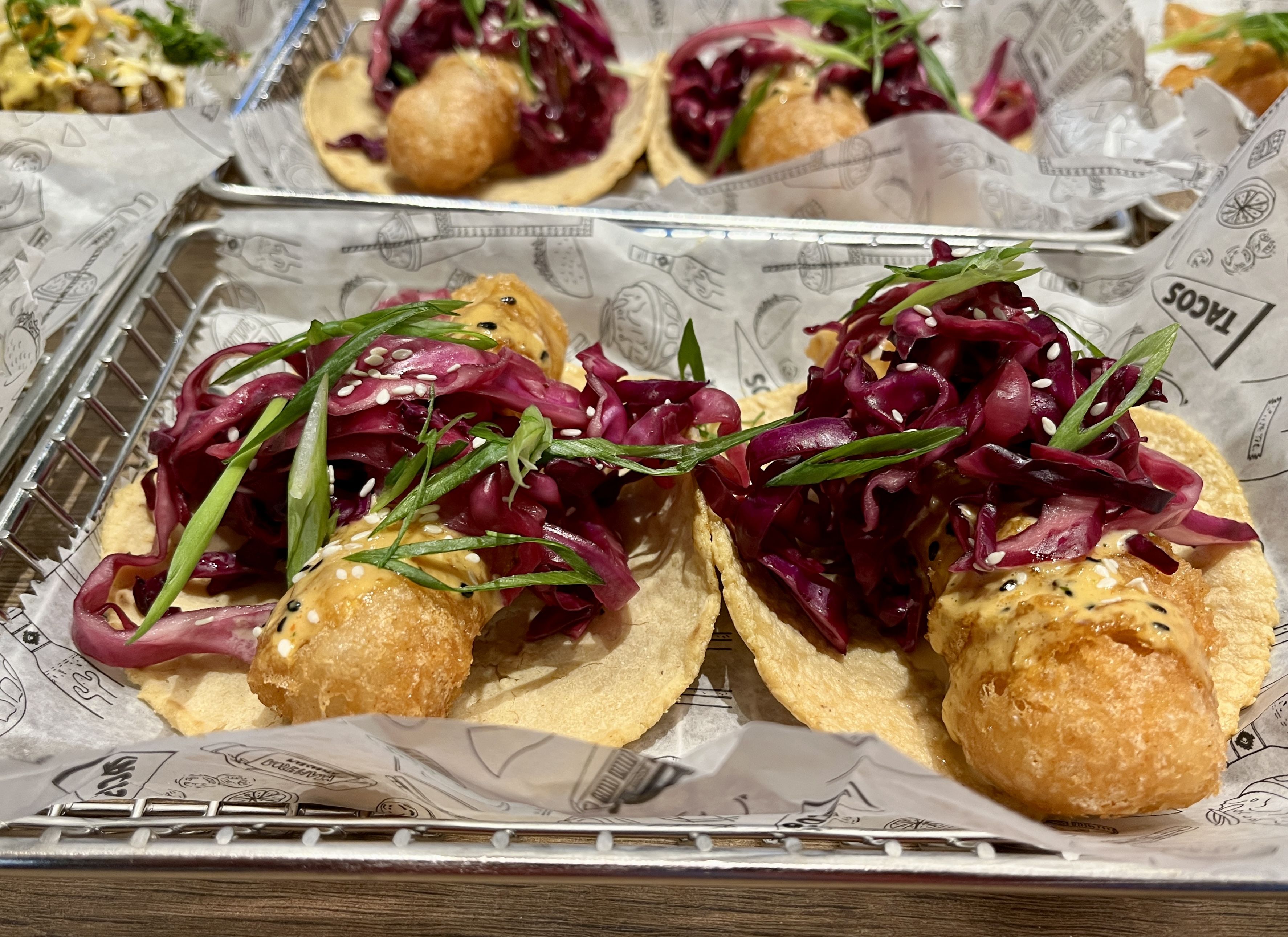 Two fried fish tacos topped with purple cabbage slaw, green onion slices, sesame seeds, and sauce on corn tortillas in a metal tray lined with printed paper.