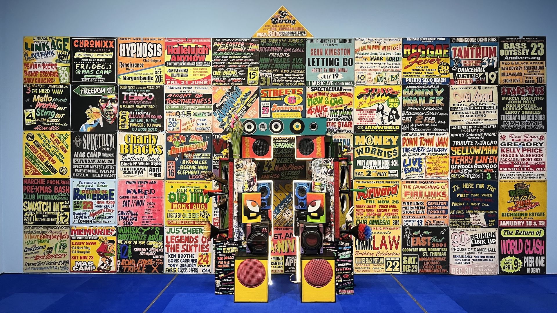 Wall covered with a dense collage of colorful concert posters and flyers; in front is a playful robot-like speaker sculpture made from boxes and speakers on a blue floor.