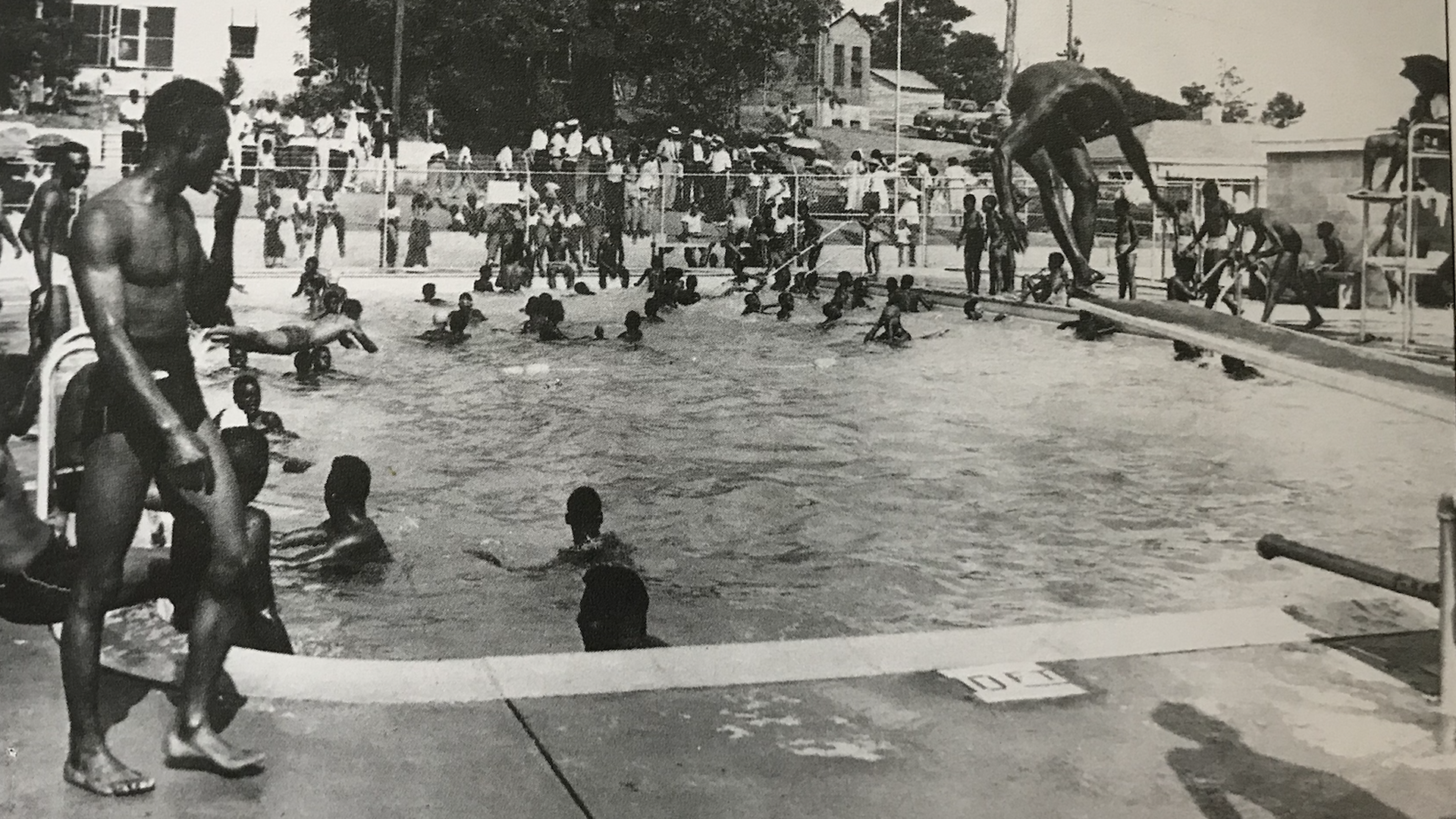 A black-and-white photo of Black swim at a public pool and a person performs a back flip into the water