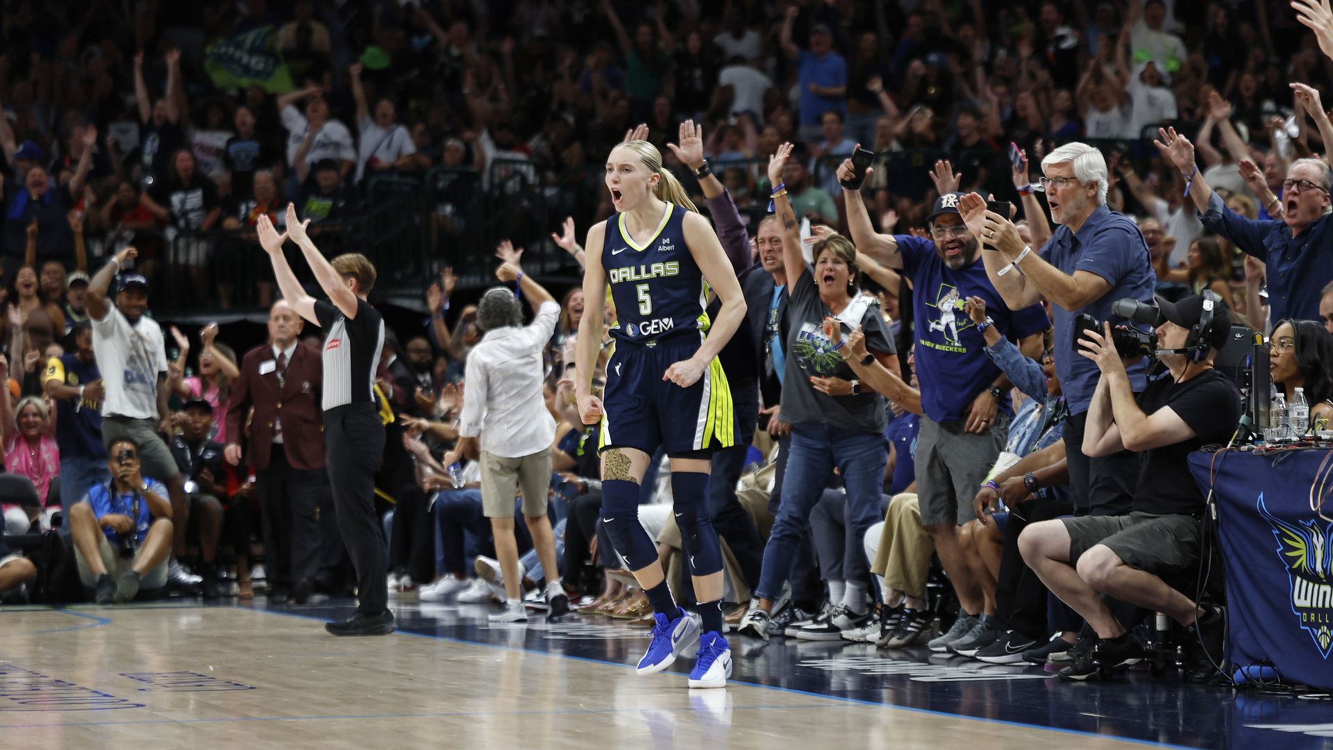 A female basketball player stands at the end of the court with dozens of people behind her, cheering