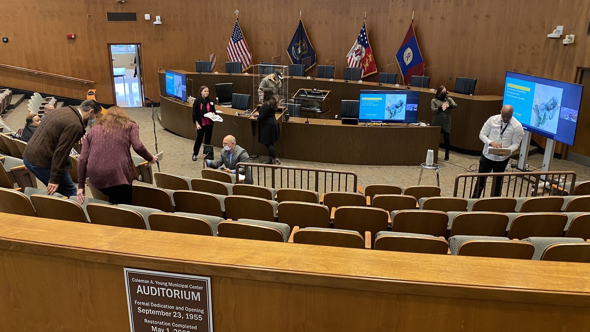 An auditorium is shown with seats and wood paneling and flags lining the back.
