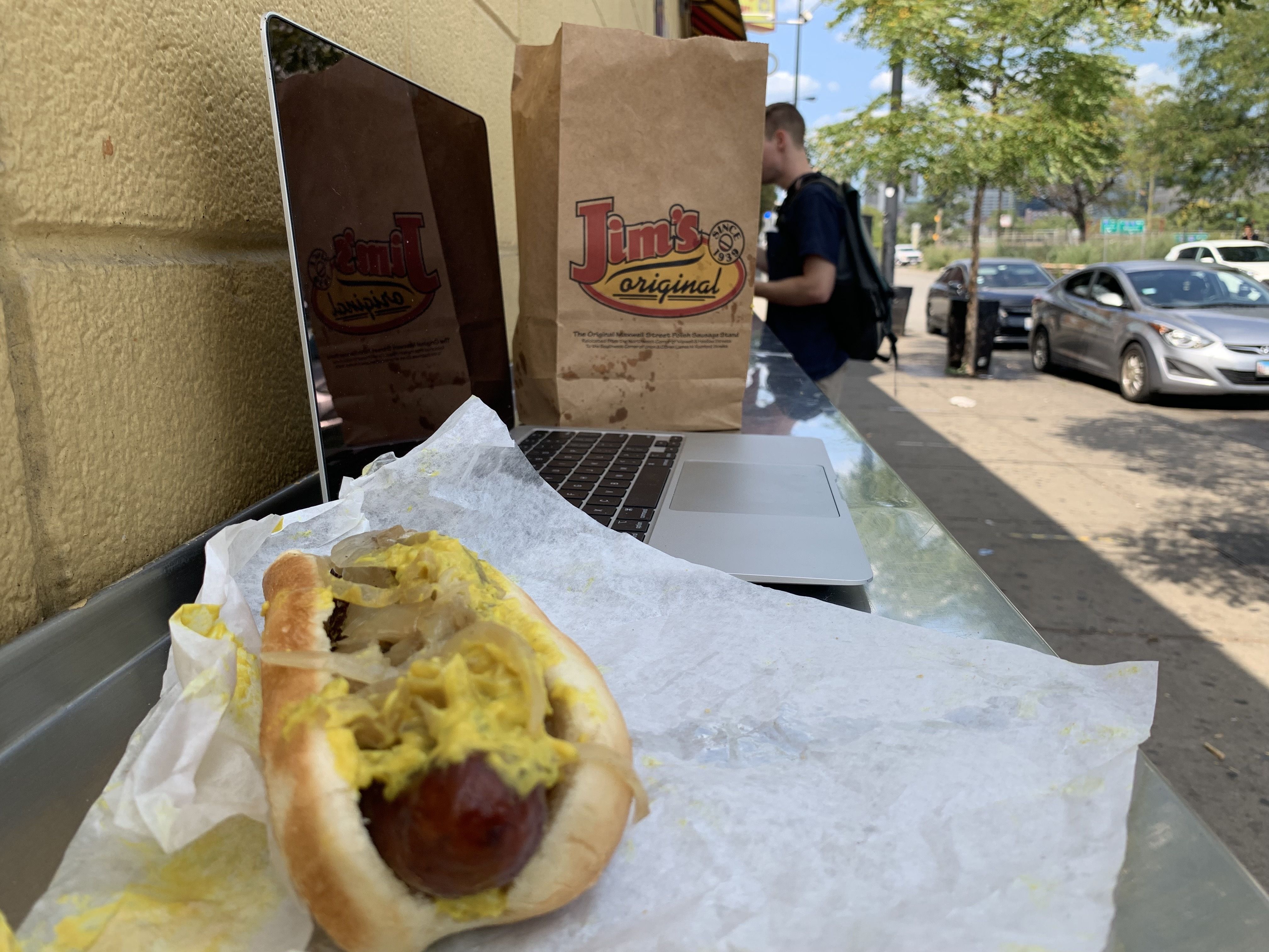 Street-side scene: laptop on a counter, a brown bag labeled "Jim's Original," and a mustard-drenched hot dog in the foreground. A backpacked person and parked cars along a tree-lined street in the background.