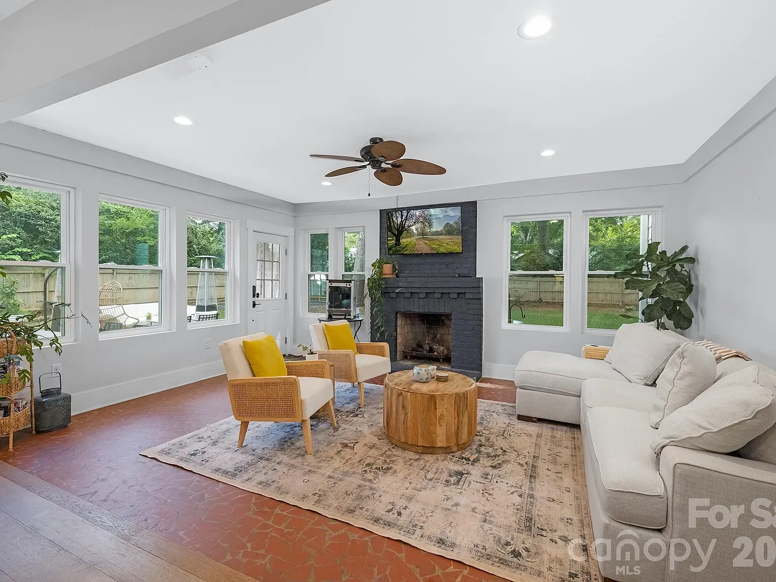 Living room with white walls, a dark brick fireplace and TV, a ceiling fan, large windows, beige sectional sofa, cream chairs with yellow cushions, round coffee table, and plants.