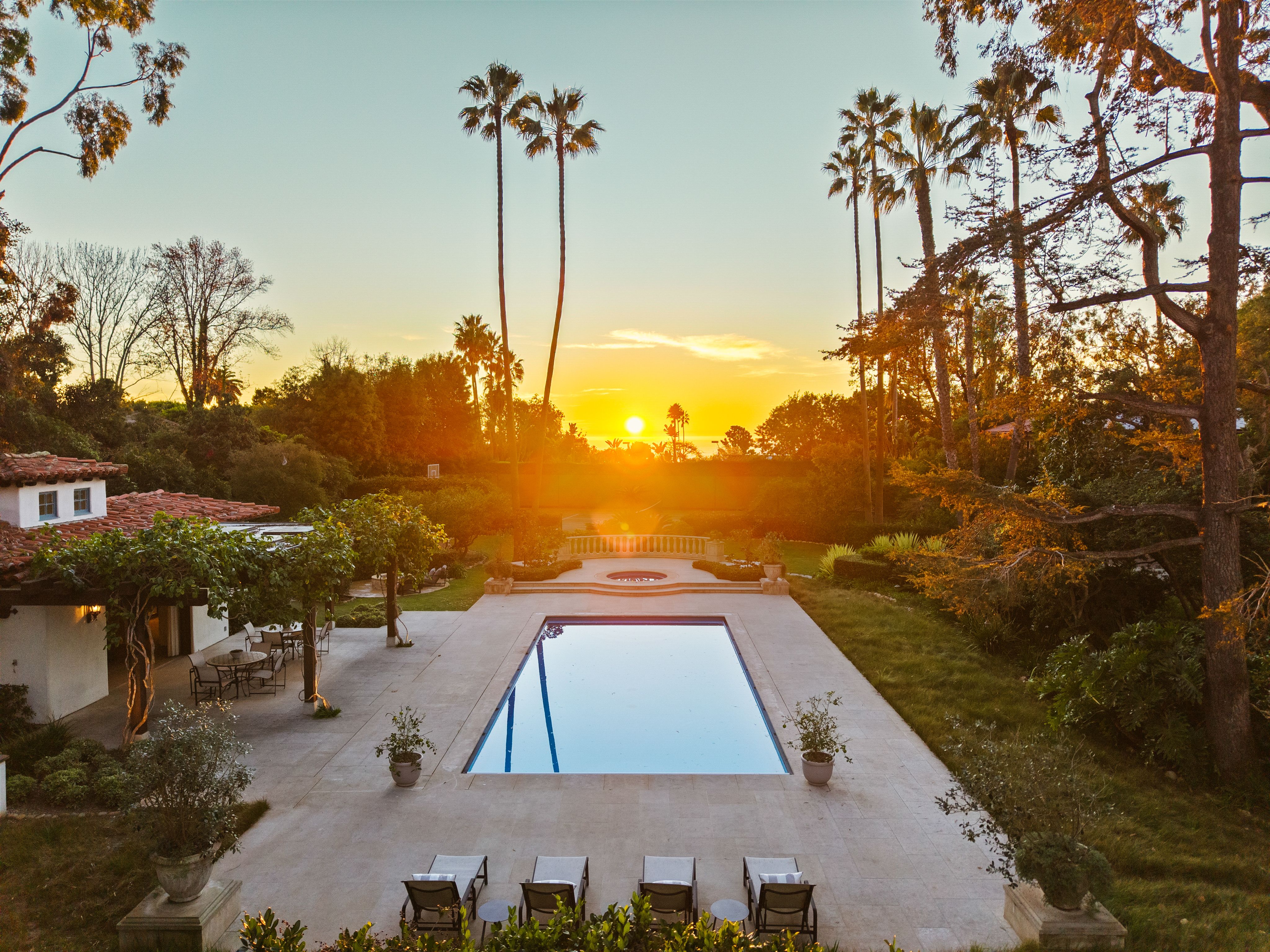 Sunset over a long rectangular pool in a courtyard, framed by tall palm trees. Warm orange light bathes the tiled patio, with lounge chairs and lush greenery around a white building on the left.