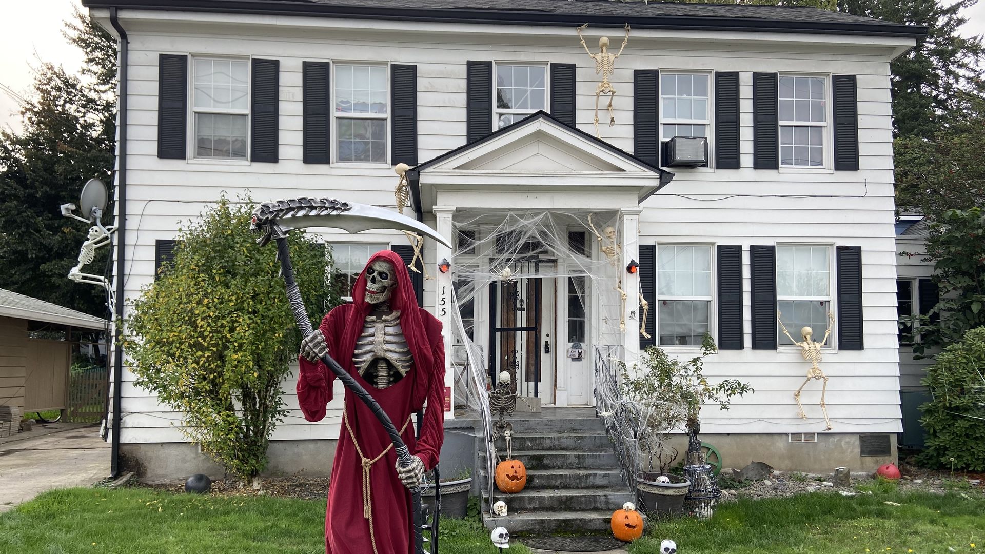 A fake skeleton wearing a red robe and holding a scythe in front of a square white house with two pumpkins on the front steps.