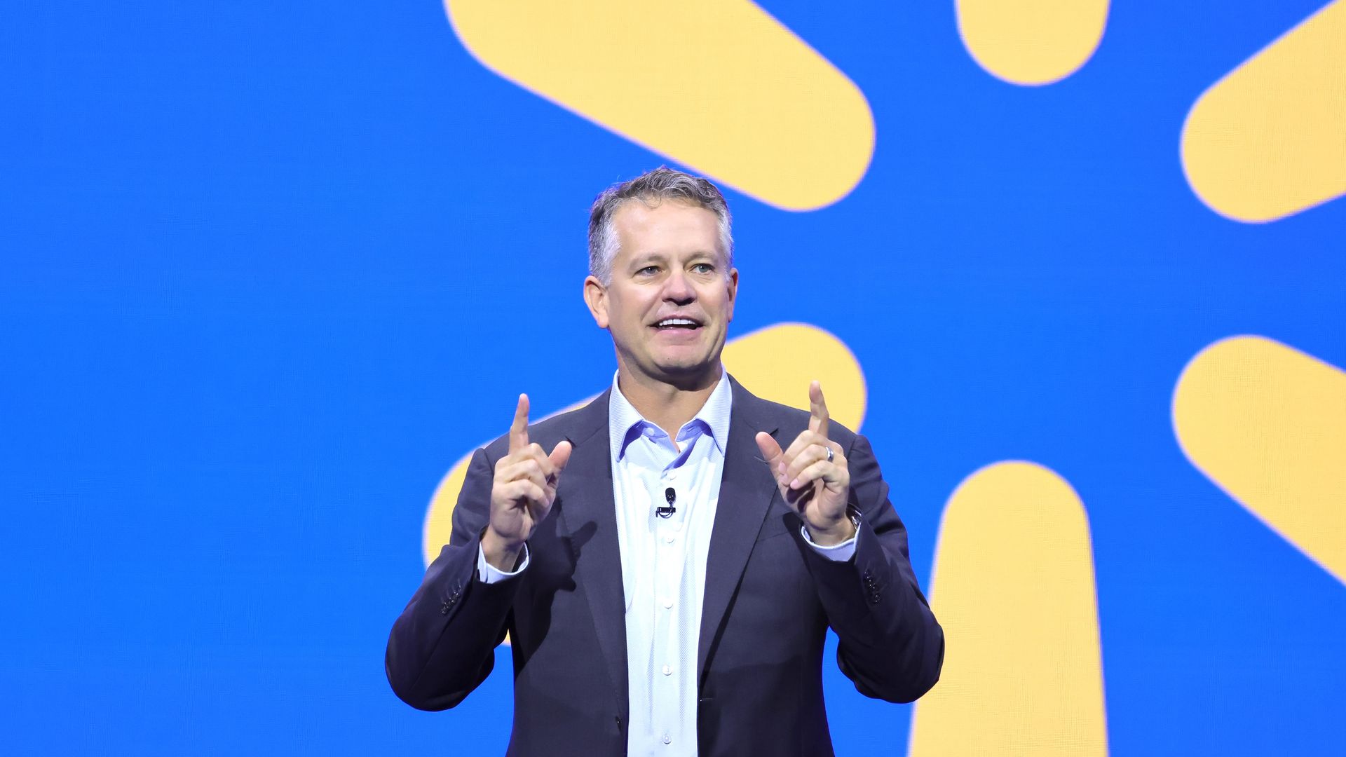 A middle-aged man in a dark blazer and light shirt speaks on stage with a lavalier mic, gesturing with both hands. A blue backdrop with large yellow abstract shapes fills the background.