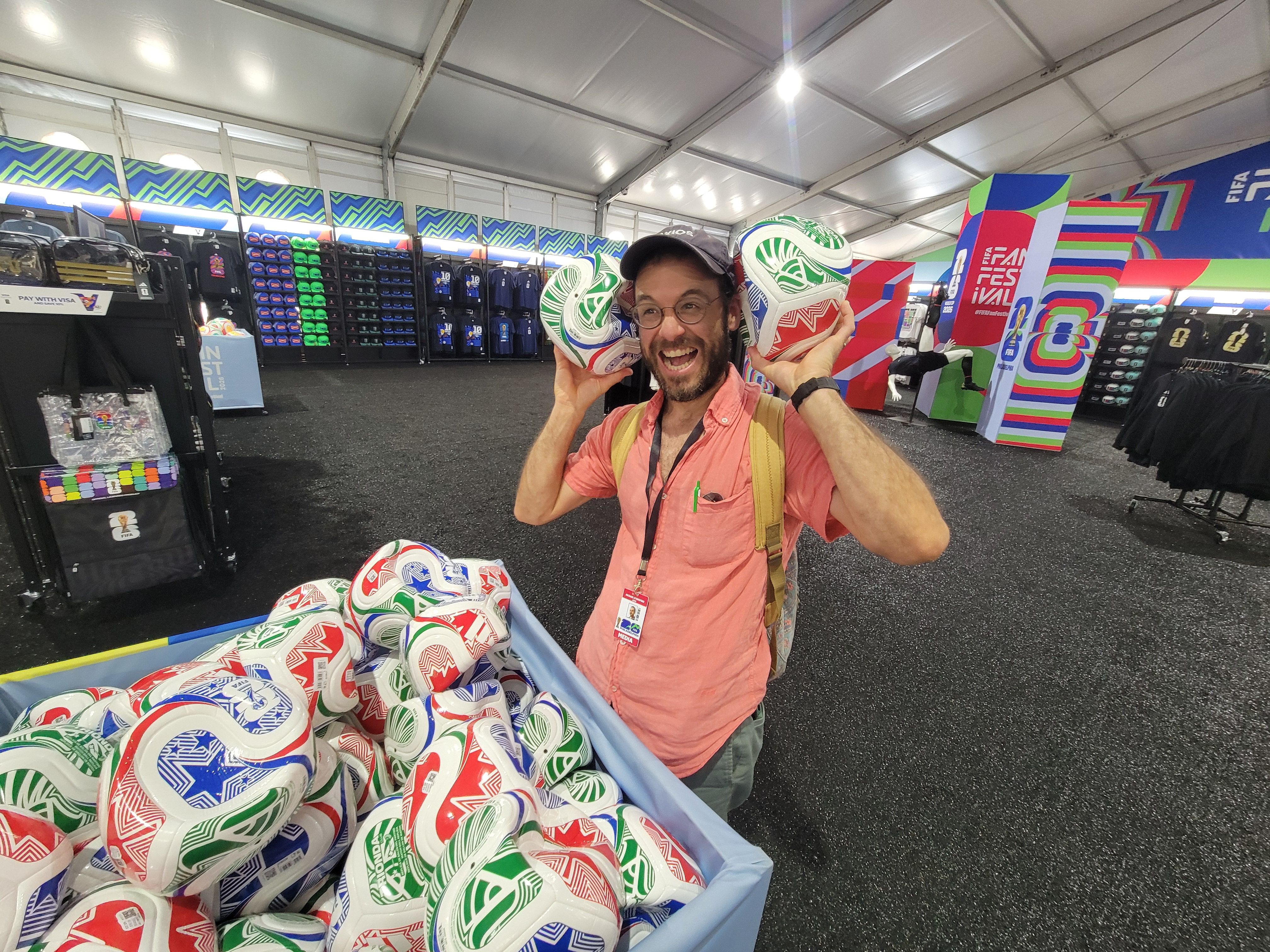 Axios reporter Mike D'Onofrio in a cap in a pink shirt stands inside a bright festival merchandise tent, holding two colorful soccer balls to his ears.