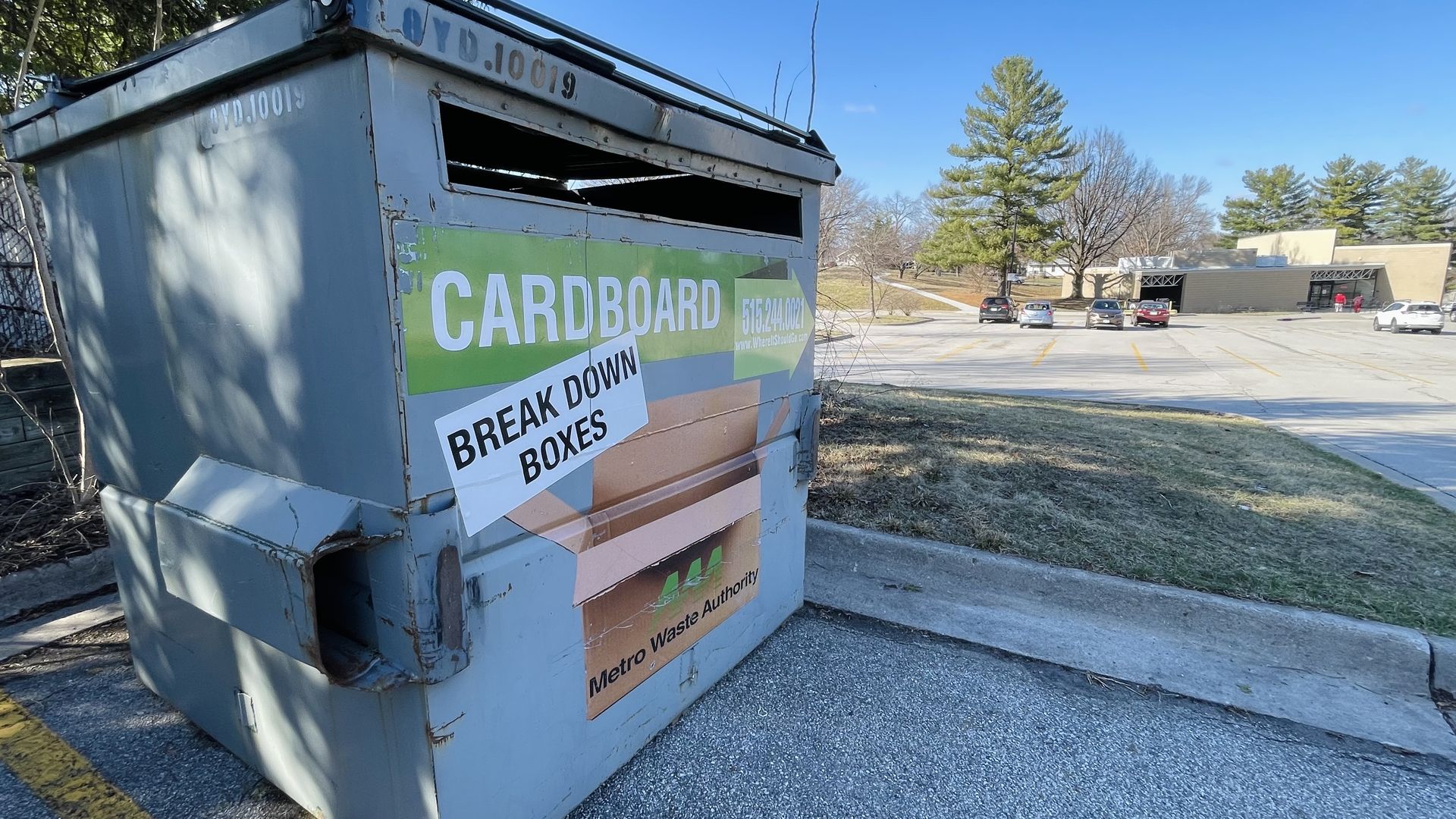 Weathered gray dumpster in a parking lot with a bright green sign that reads "CARDBOARD" and a taped "BREAK DOWN BOXES" notice. Cars and a building are in the background under a blue sky.