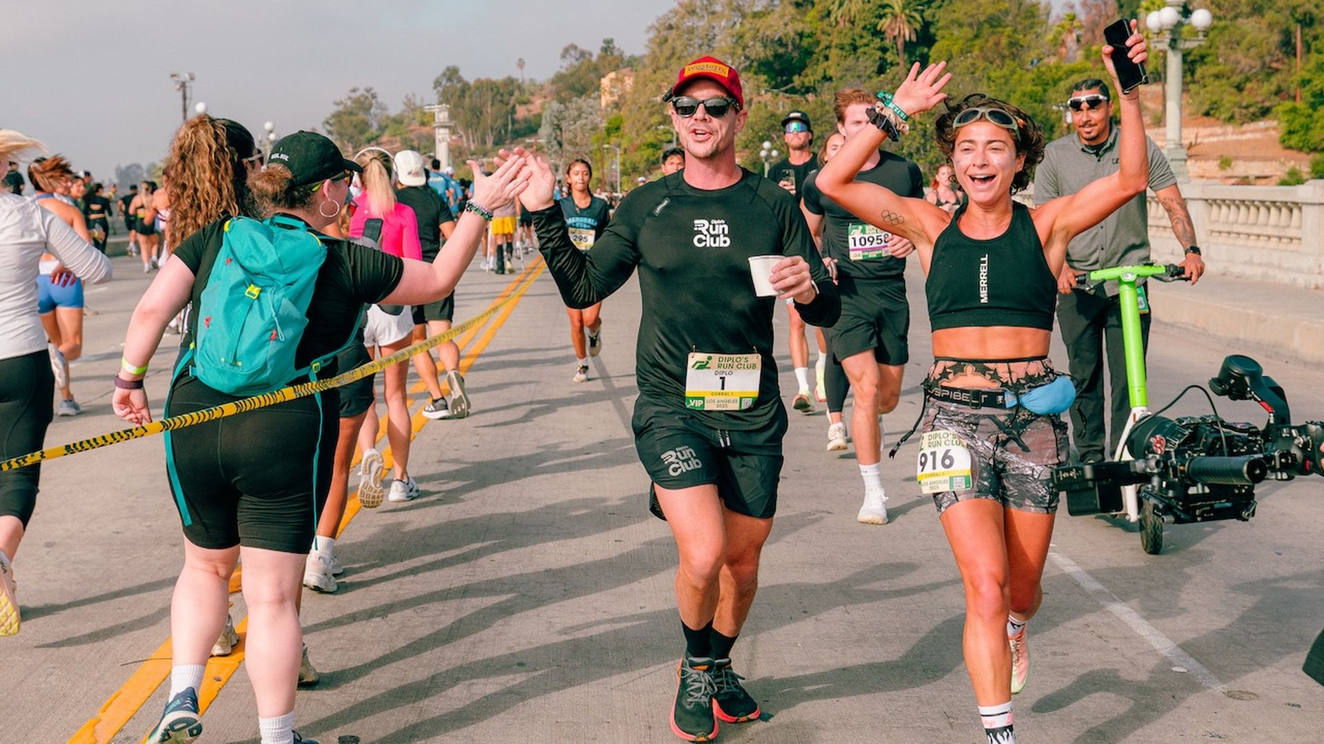 Runners high-fiving supporters along a road during a race event with trees and lampposts in the background on a sunny day.