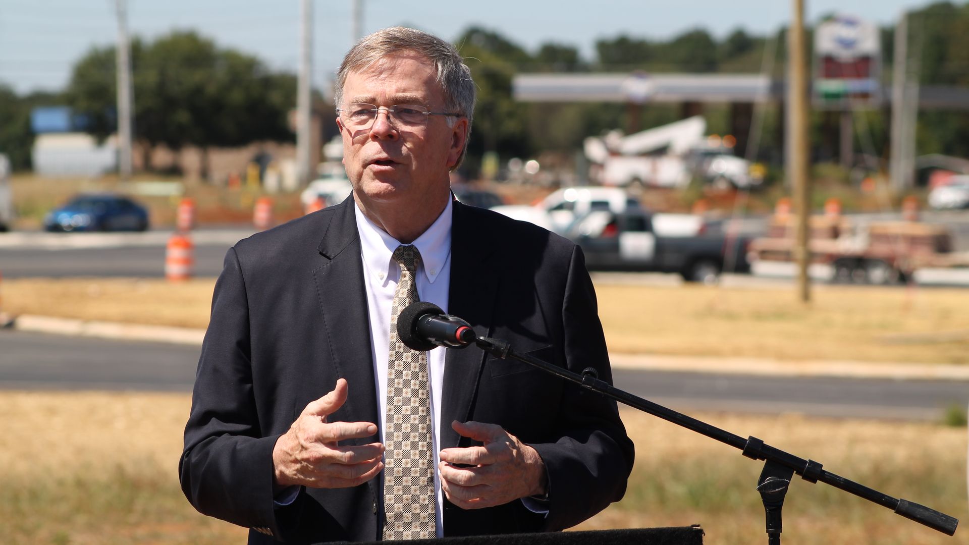 Man in dark suit and patterned tie speaking outdoors at a microphone stand, with blurred traffic and orange construction barrels in the background on a sunny day.