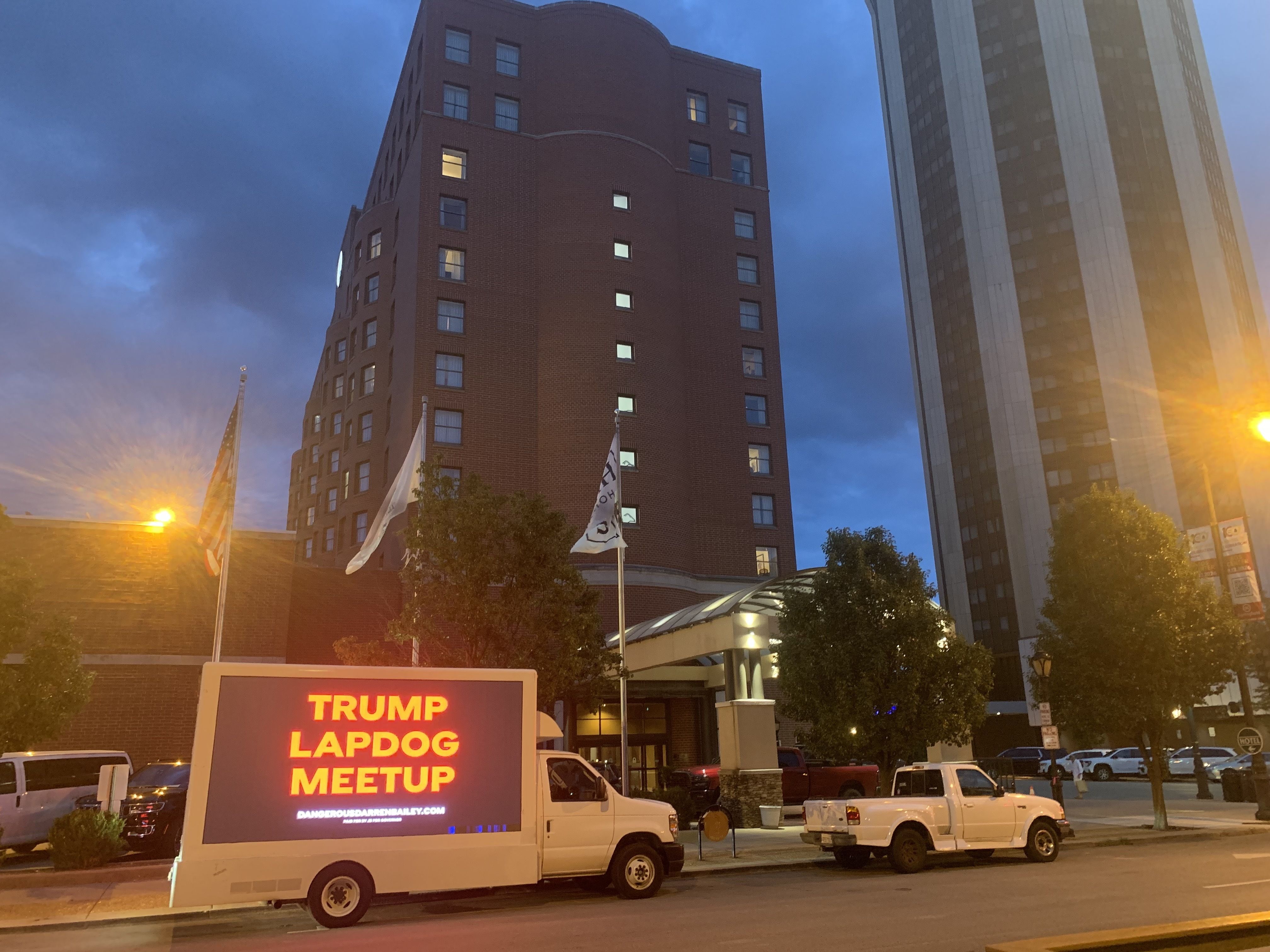 Evening urban scene: a tall curved brick building center, a glass tower to the right, blue dusk sky, flags and trees, with a white van in front showing a red LED sign reads TRUMP LAPDOG MEETUP.