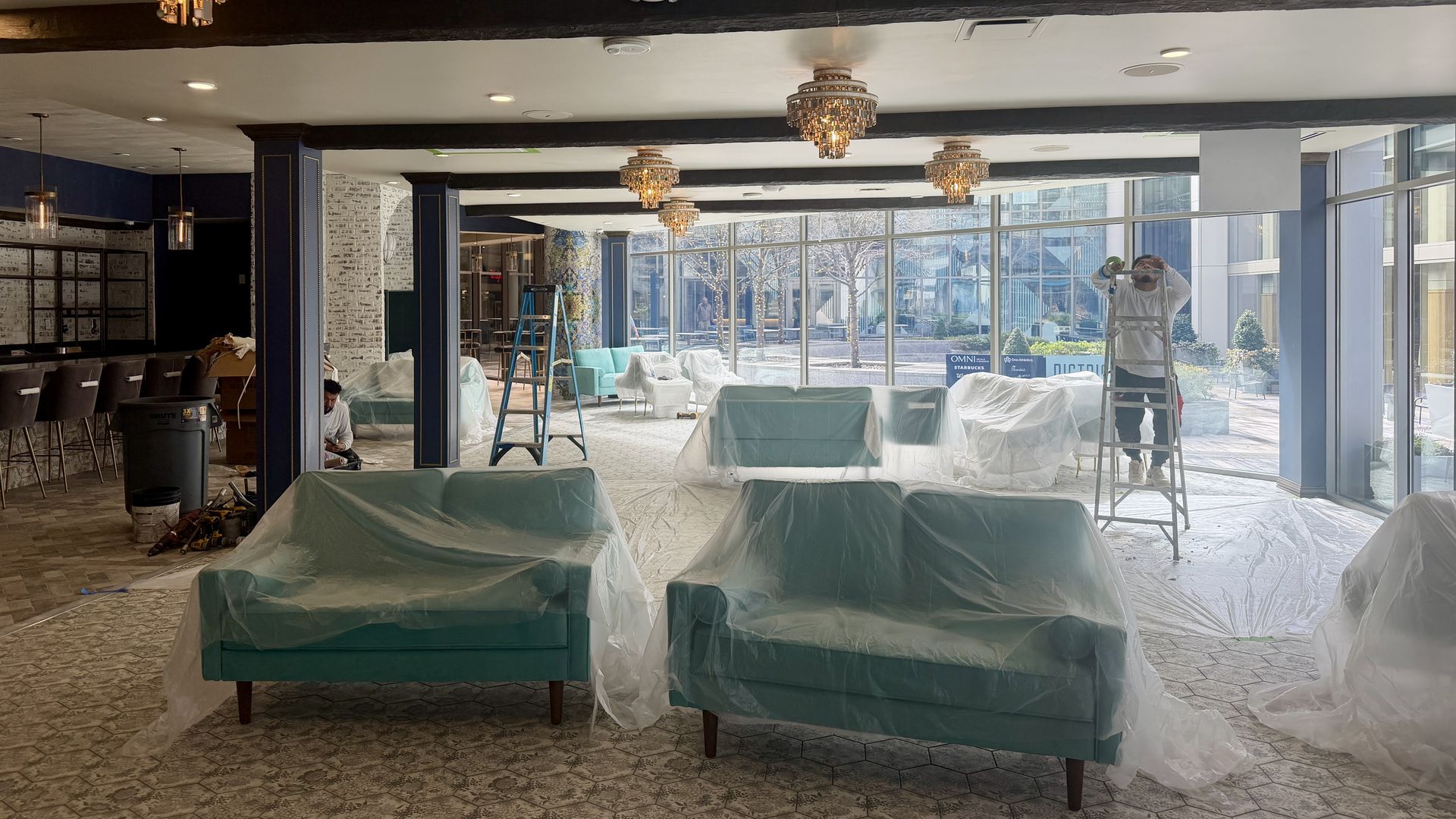 Room with teal sofas covered in plastic, chandeliers on ceiling, ladders, and workers doing maintenance near large glass windows showing outdoor cityscape.