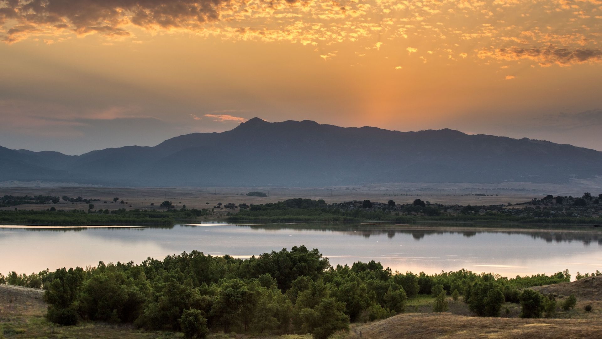 A landscape photo of a lake with trees at the forefront and a mountain in the background at sunset. 