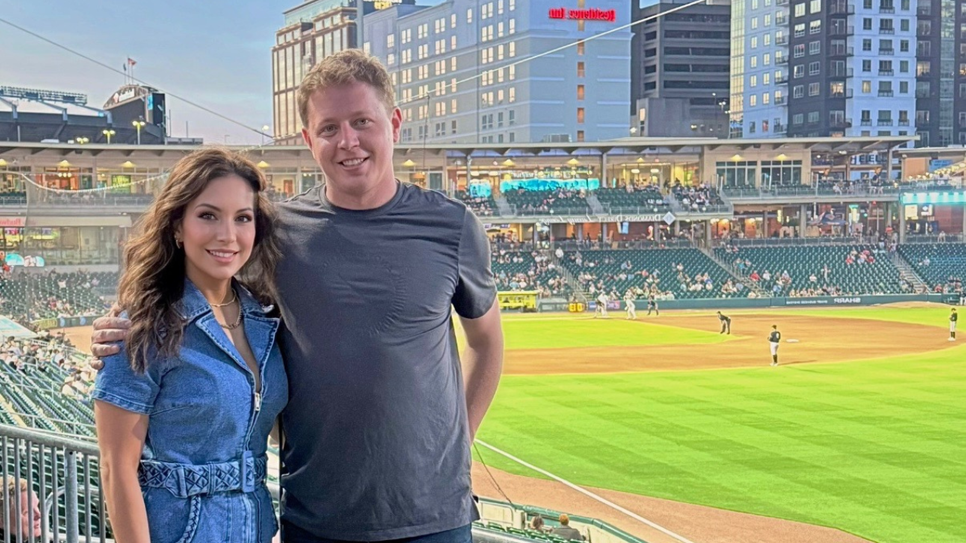 A man in a dark gray t-shirt and a woman in a blue belted denim dress smile at a baseball stadium during a game, with green field and spectators in the background.