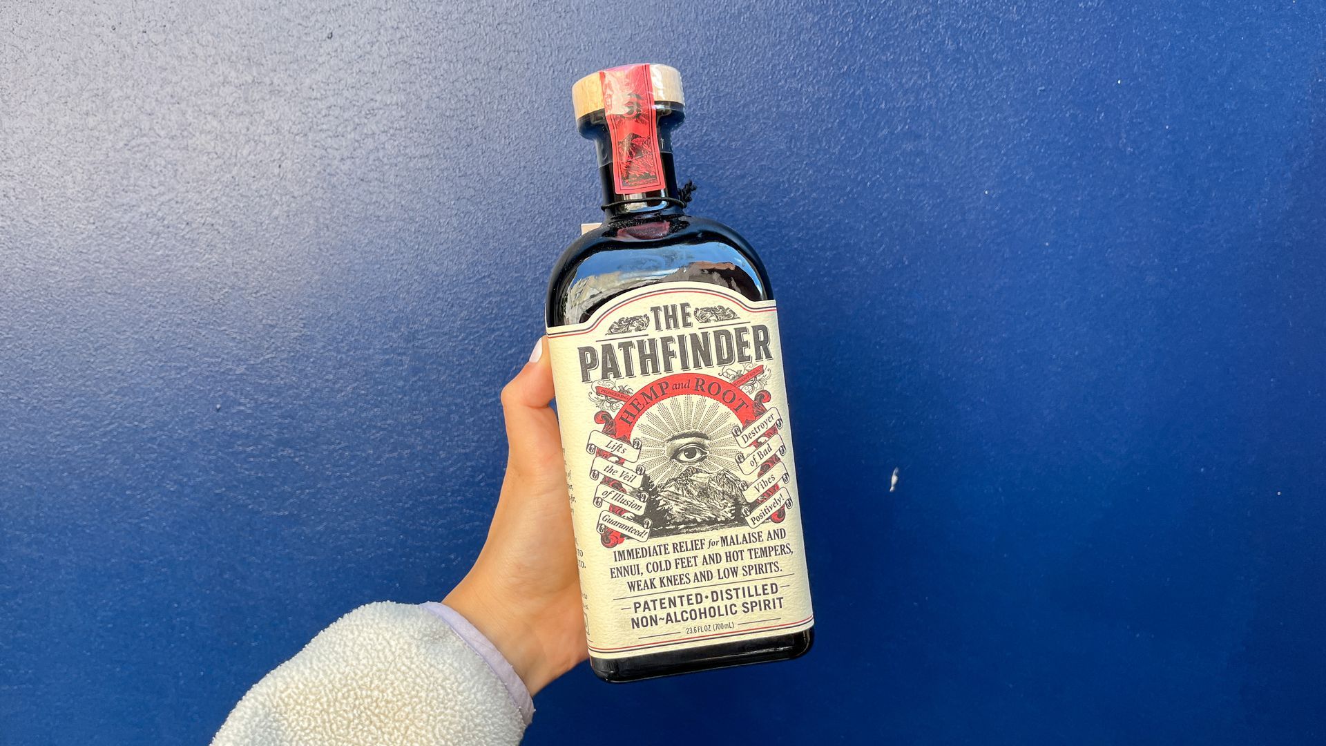 A woman's hand holds up The Pathfinder bottle in front of a blue wall.
