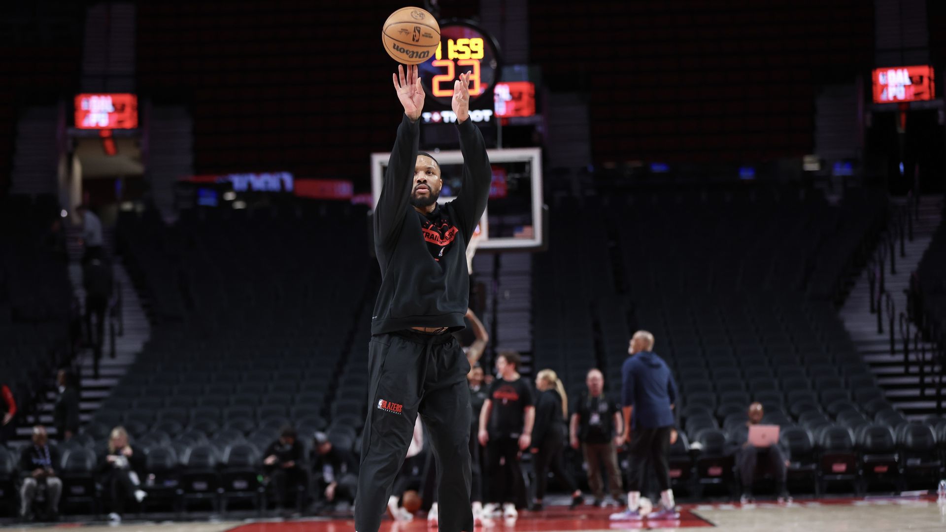 Basketball player in black Portland Trail Blazers warm-up outfit shooting a basketball on court with empty stands and several people in the background.