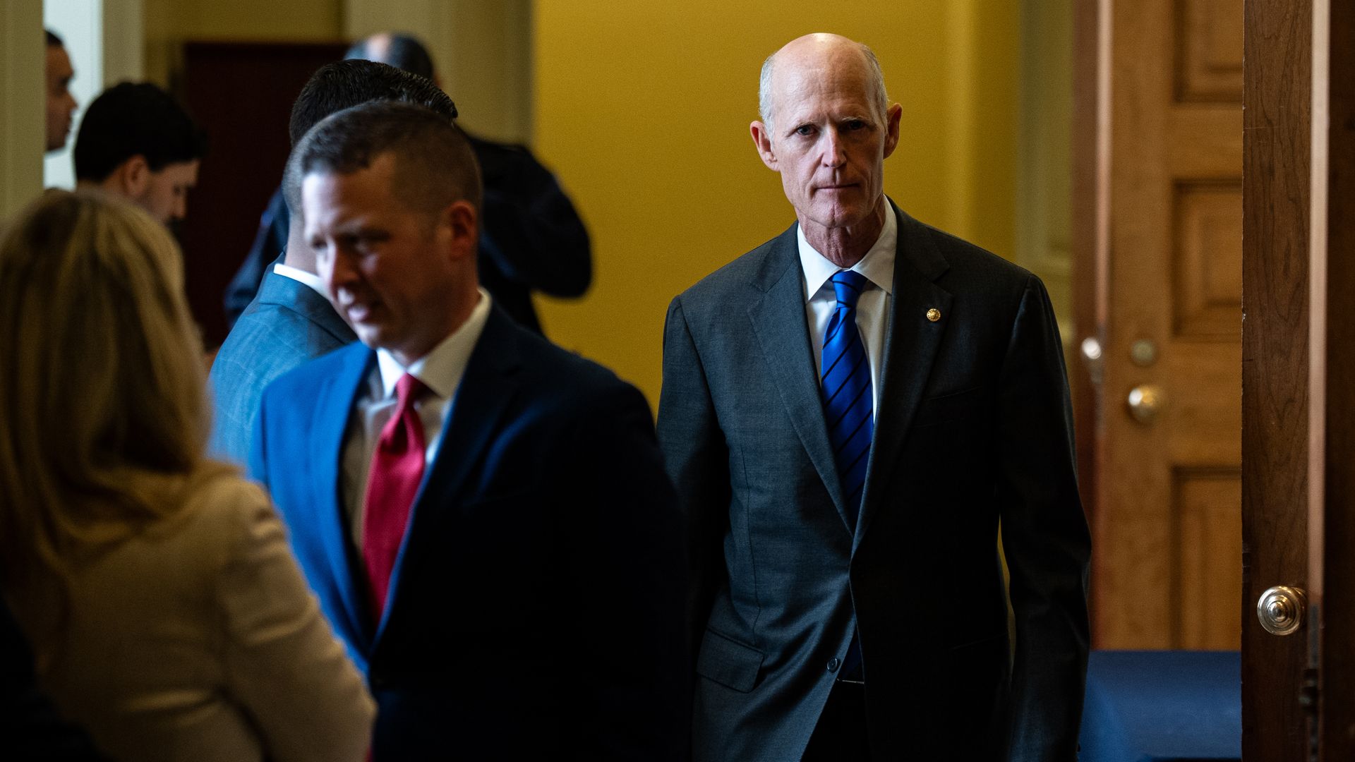 Tall man in a dark suit and blue striped tie walks through a doorway in a hallway; several suited people stand nearby as yellow walls and a wooden door frame surround the scene.
