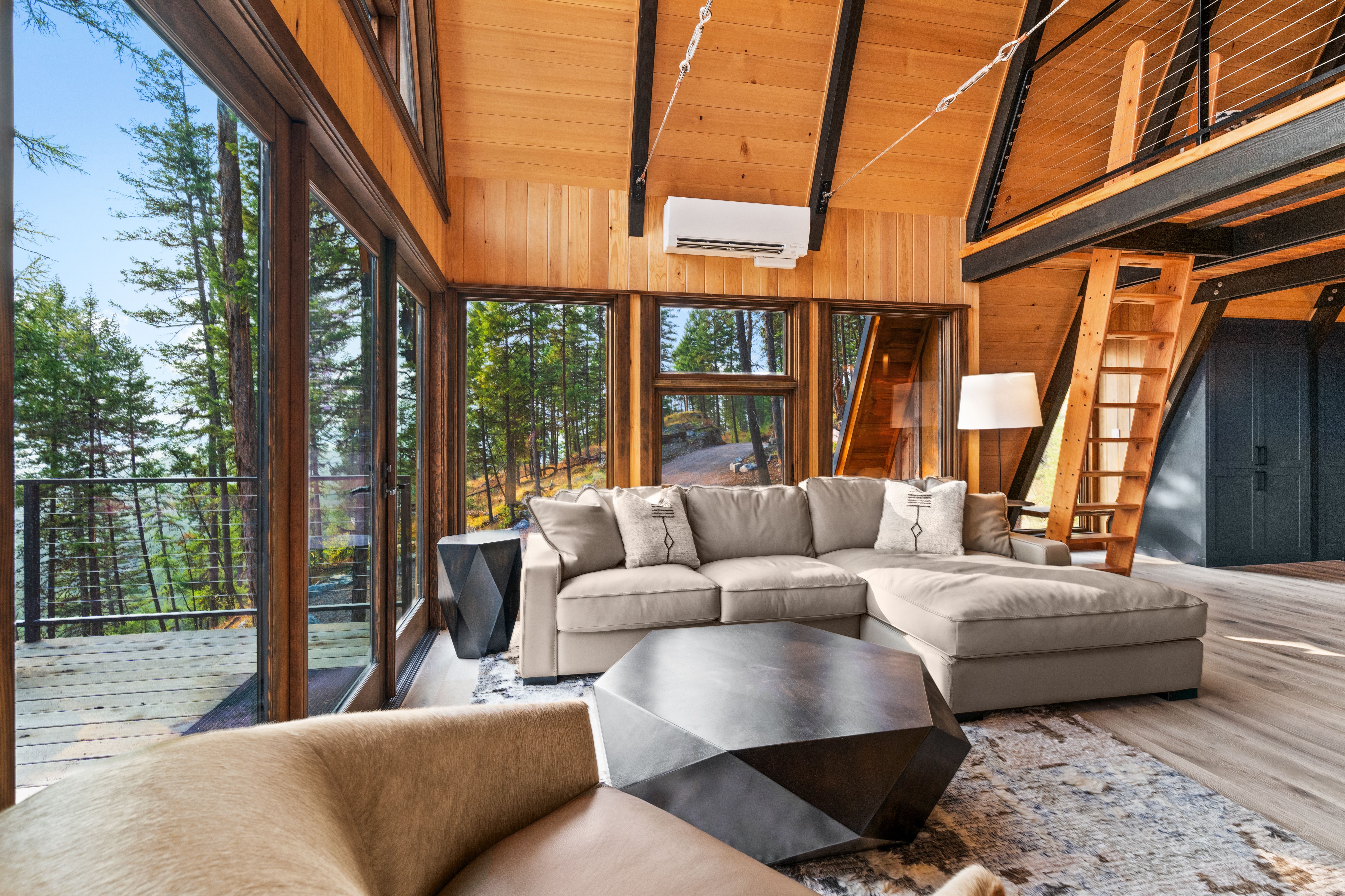 An interior view of a living room with wooden paneled walls and large windows showing a forest outside.