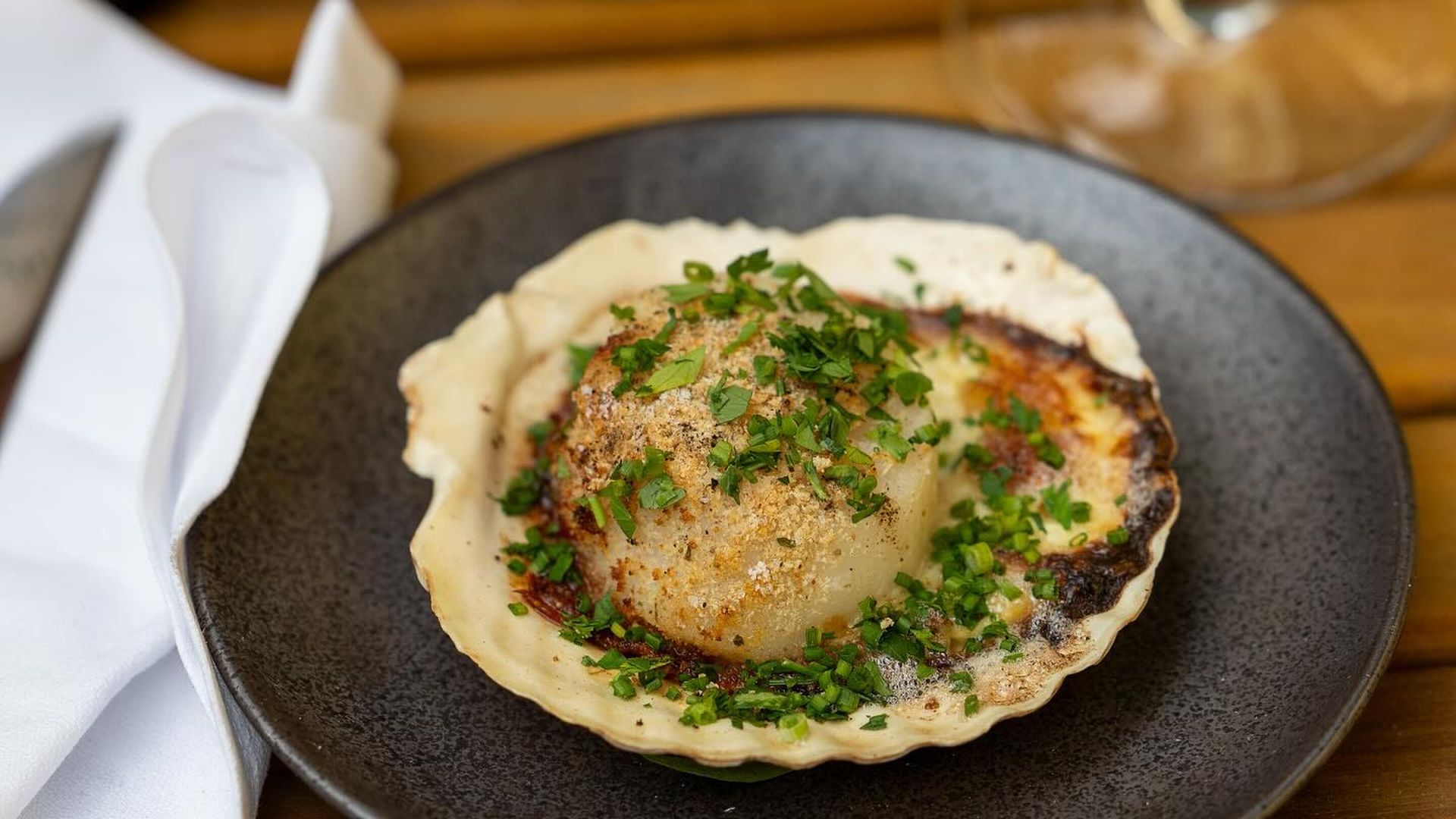 Seared scallop in a scallop shell on a dark plate, garnished with chopped parsley; blurred background shows a wine bottle, a vase of yellow flowers, and a glass of white wine.