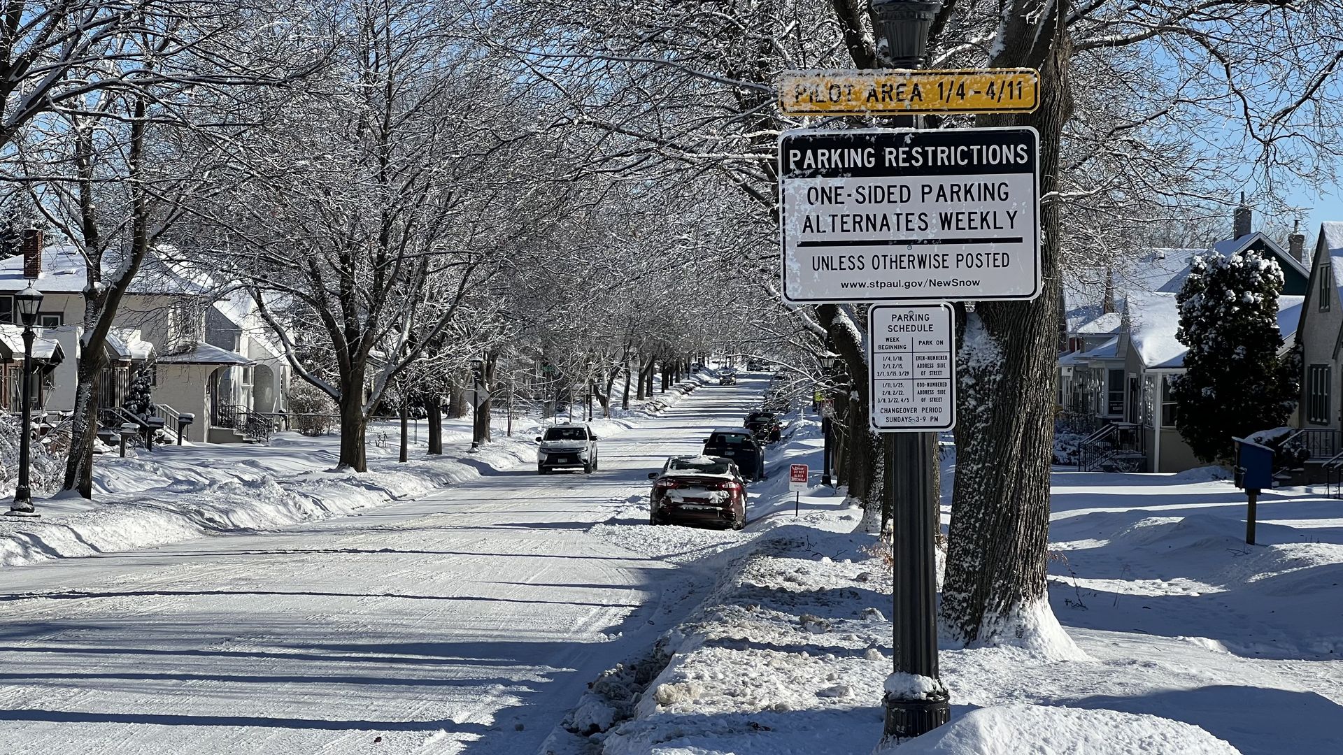 Snow-covered residential street with bare trees, parked cars, and a streetlamp displaying parking restriction signs under a clear blue sky.
