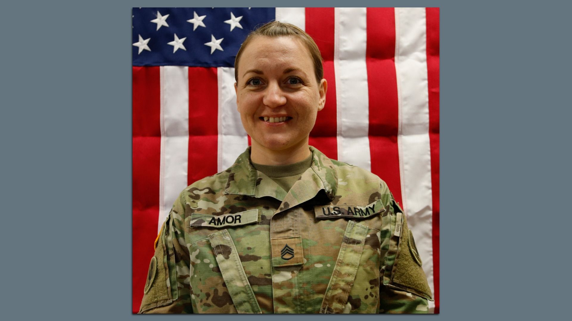 U.S. Army soldier Nicole Amor in a camouflage uniform stands in front of a U.S. flag with red and white stripes and white stars on a blue field.