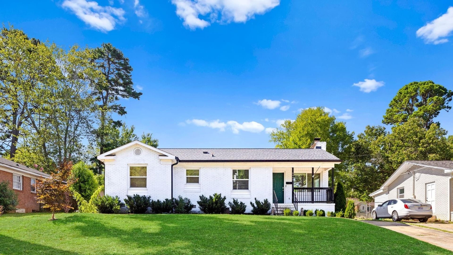 Single-story white brick house with green front door and black porch railing, surrounded by green lawn, trees, and blue sky, with a white car parked in driveway on the right.
