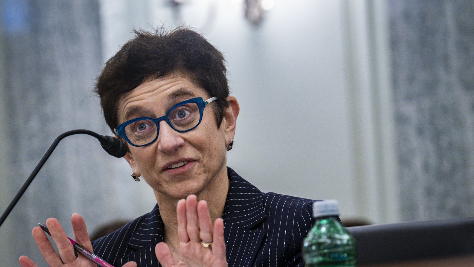 A women with short hair and blue glasses holds up her palms while speaking. She is seated.