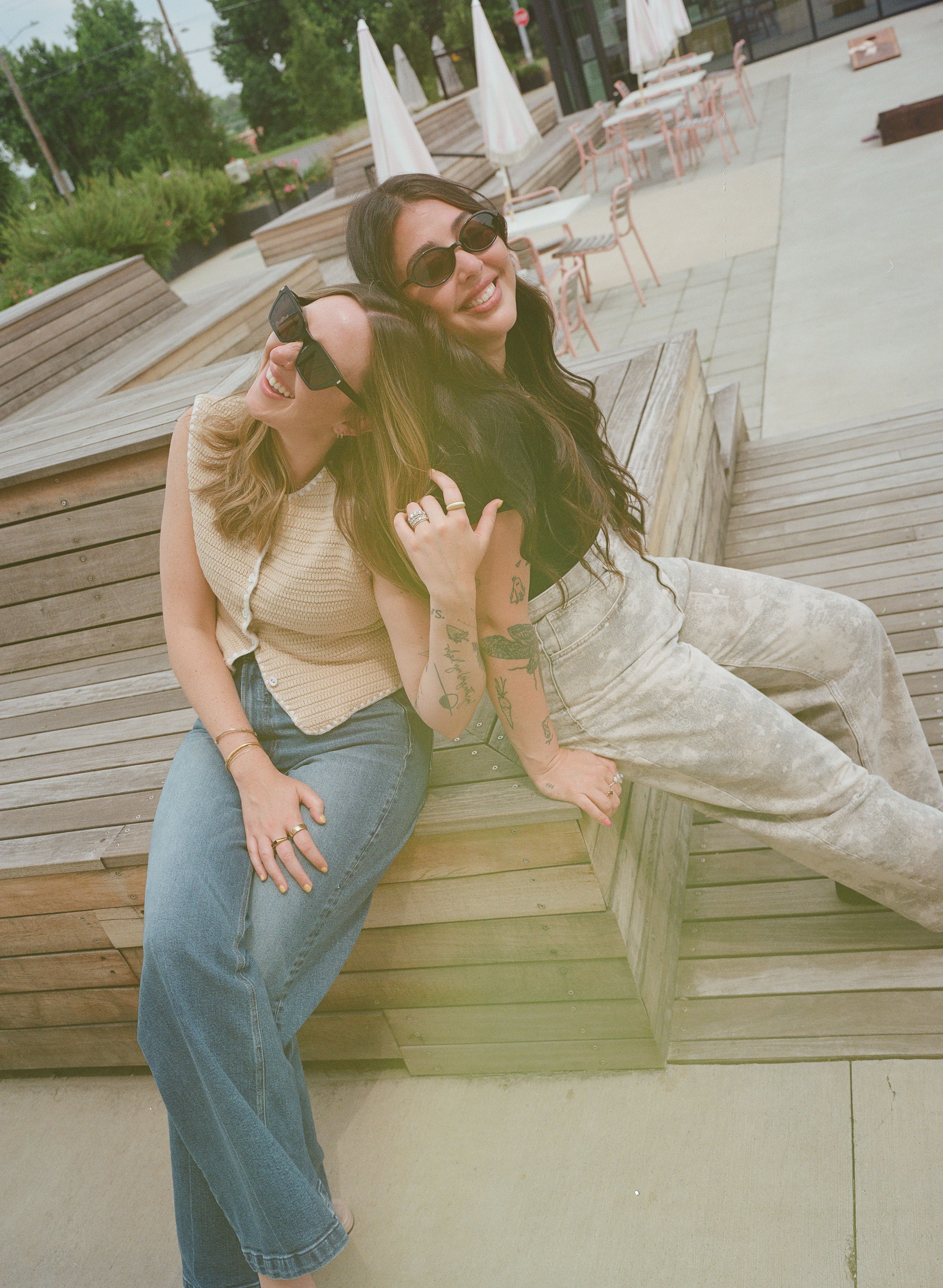 Two friends wearing sunglasses sit close on wooden steps outdoors, smiling. One in a beige sleeveless knit top and blue jeans; the other in light denim with tattoos. Pink chairs and white umbrellas nearby.