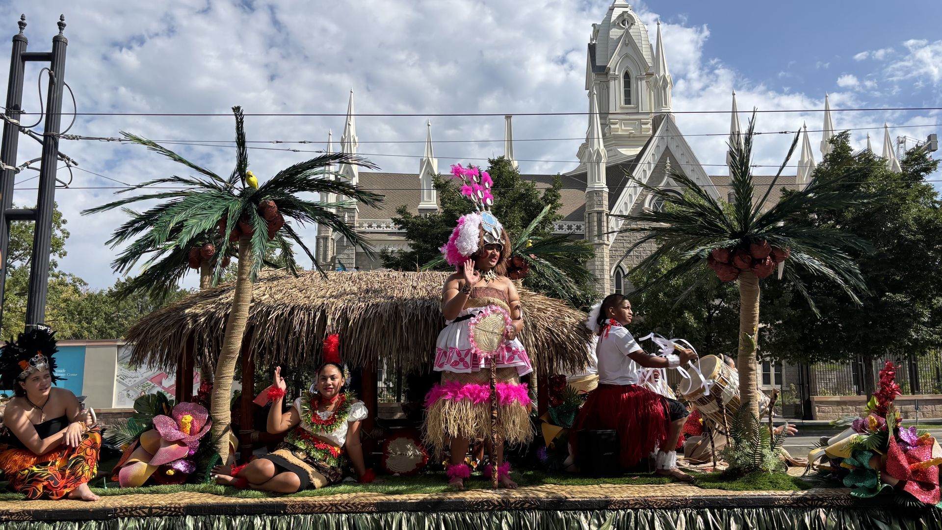 People in colorful traditional Polynesian attire on a float with fake palm trees and hut, set against a blue sky and church building in the background.