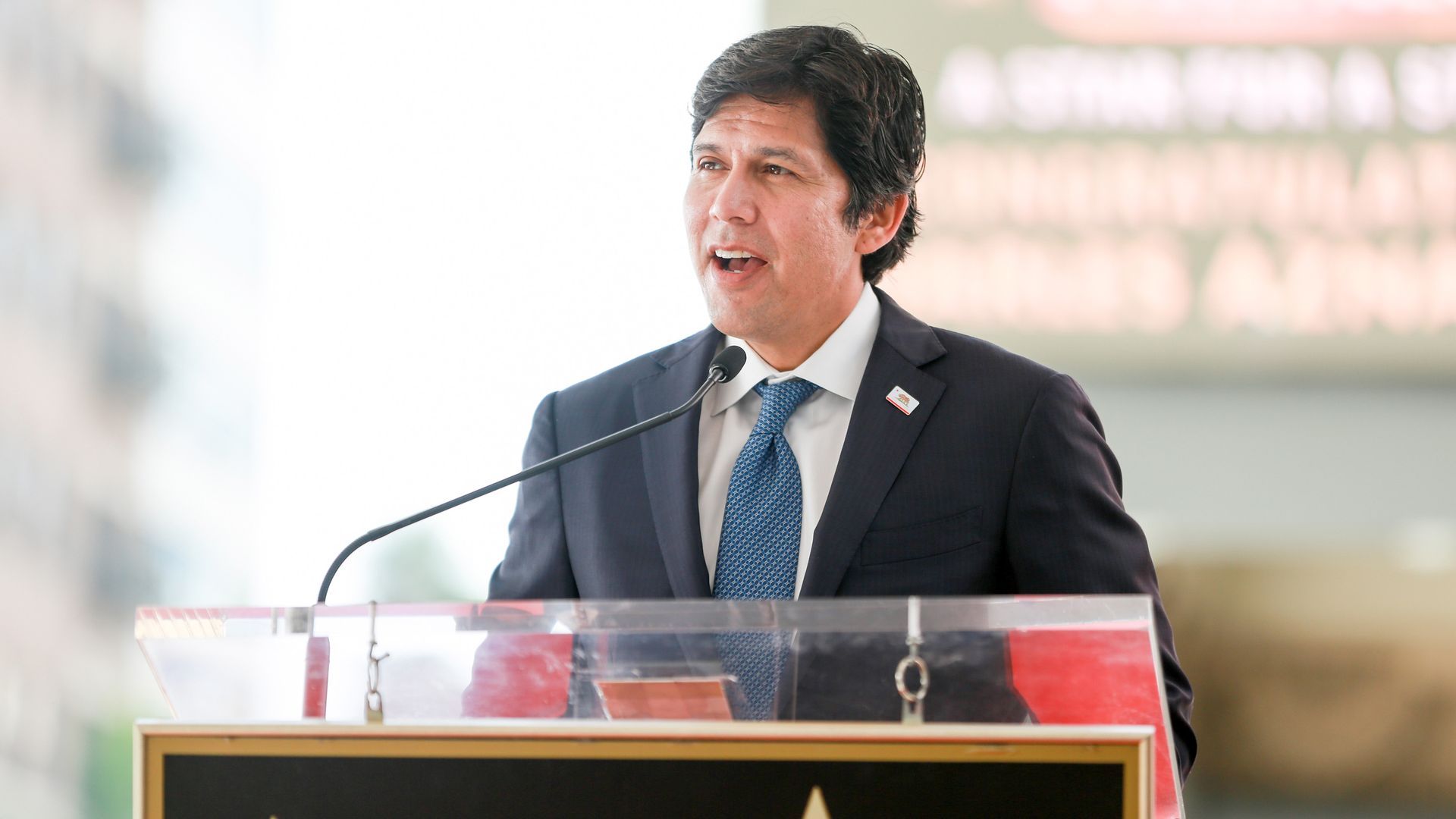 Kevin de León wearing a suit and speaking from a podium