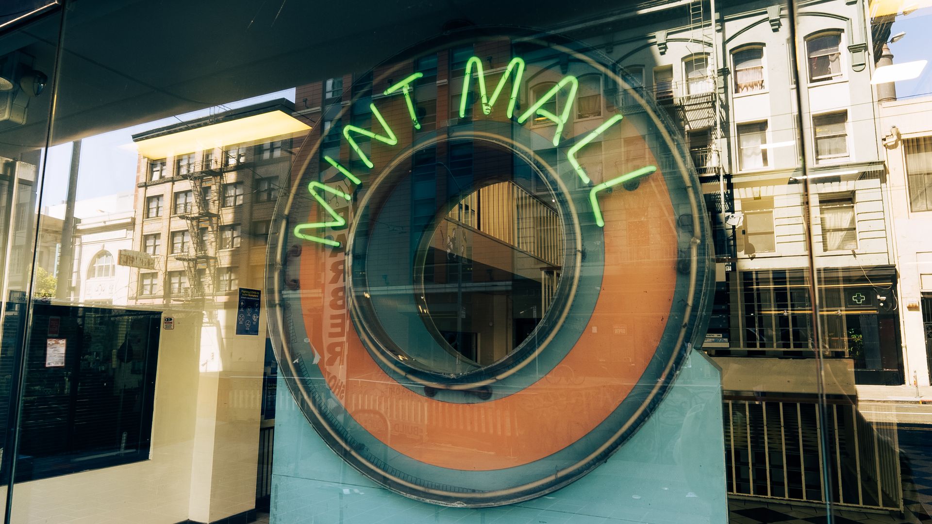 Reflection of urban buildings in a large glass window displaying a neon green "MINT MALL" sign around a circular orange and green design.