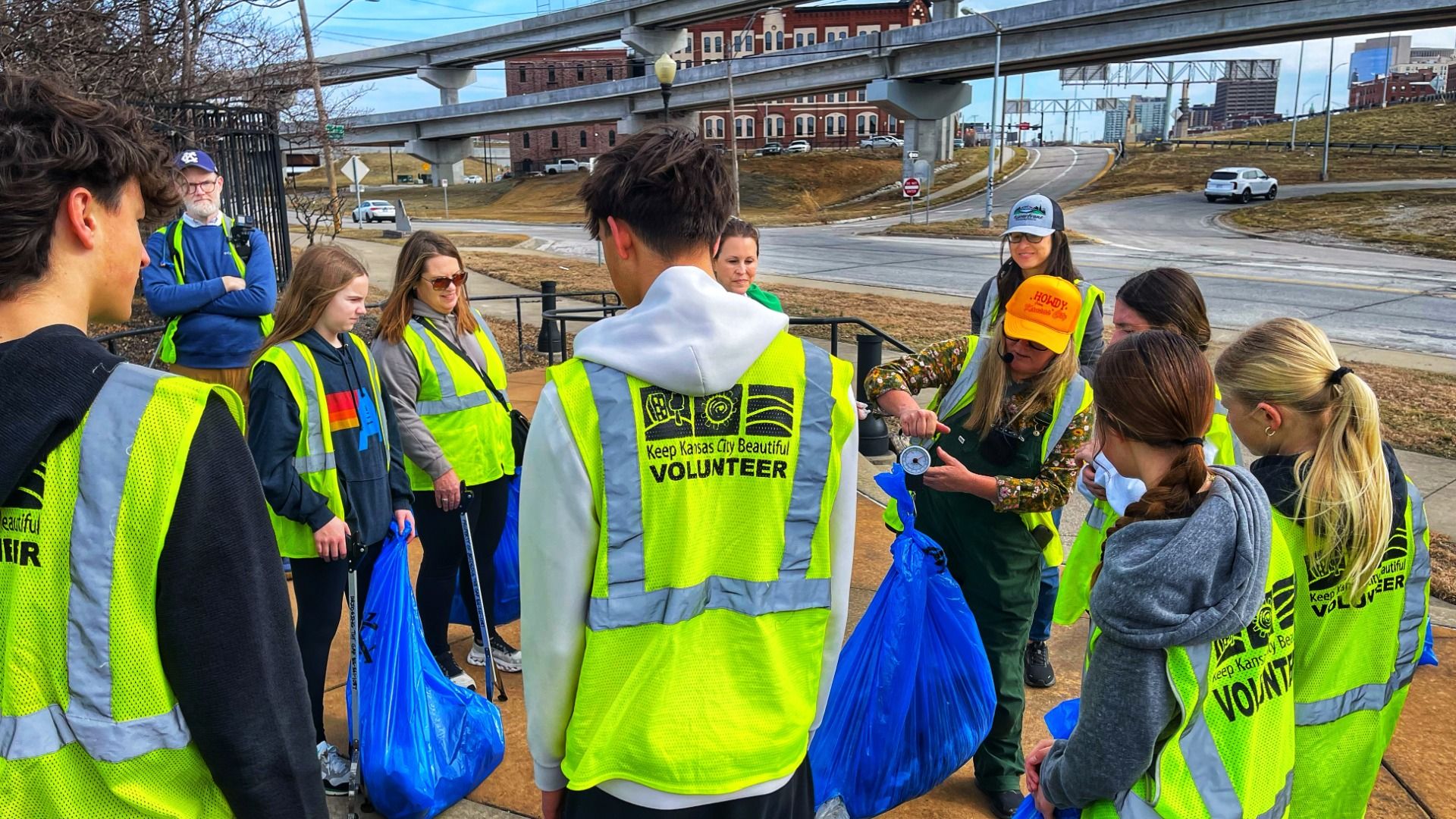 Group of volunteers in bright yellow safety vests gather on a sidewalk with blue trash bags; one person demonstrates a tool while urban overpasses and brick buildings loom in the background.