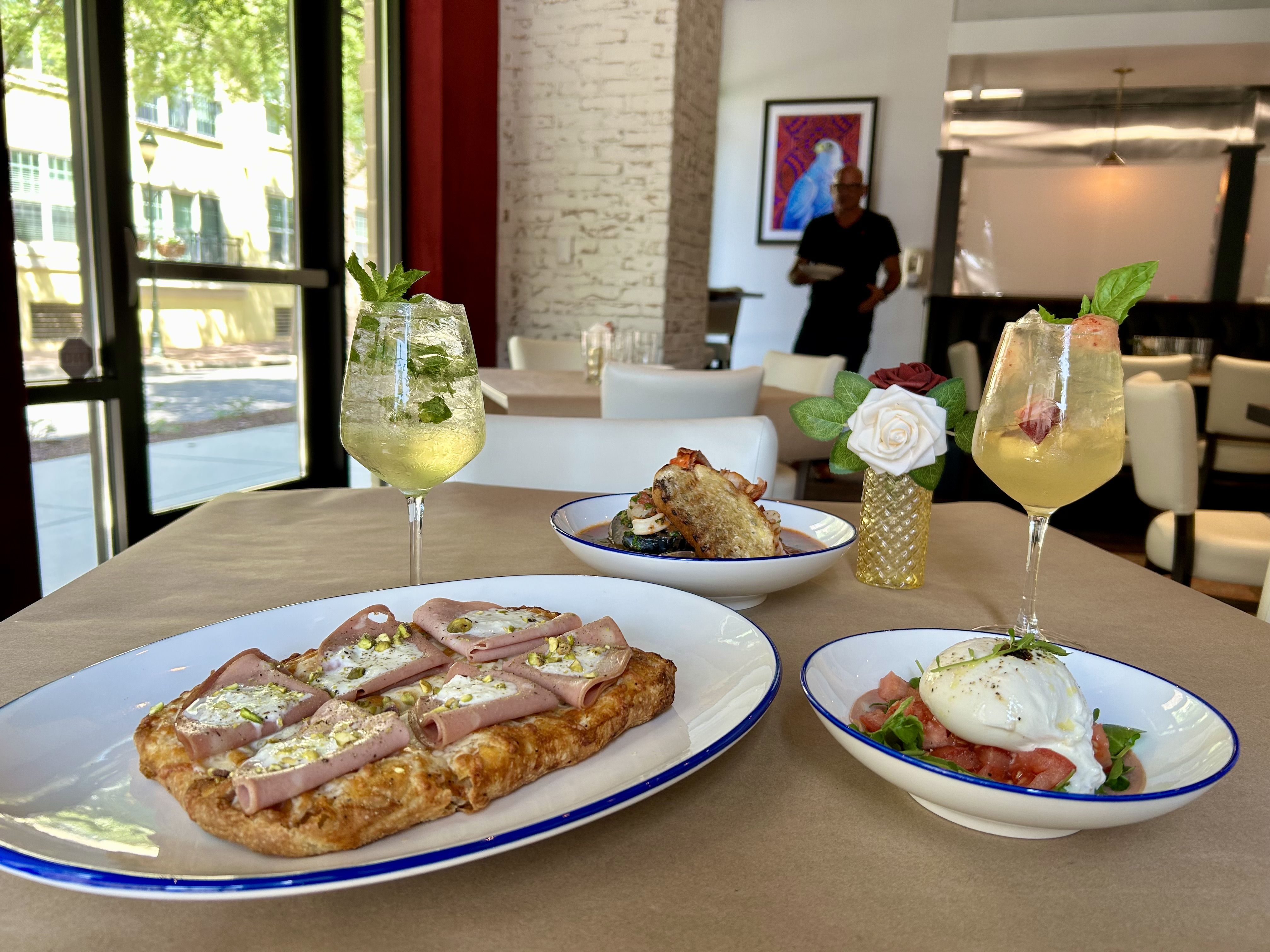 Table with two light yellow cocktails garnished with mint, a flatbread topped with ham and cheese, a dish of burrata with tomatoes, another bowl with bread and seafood in a cozy restaurant.