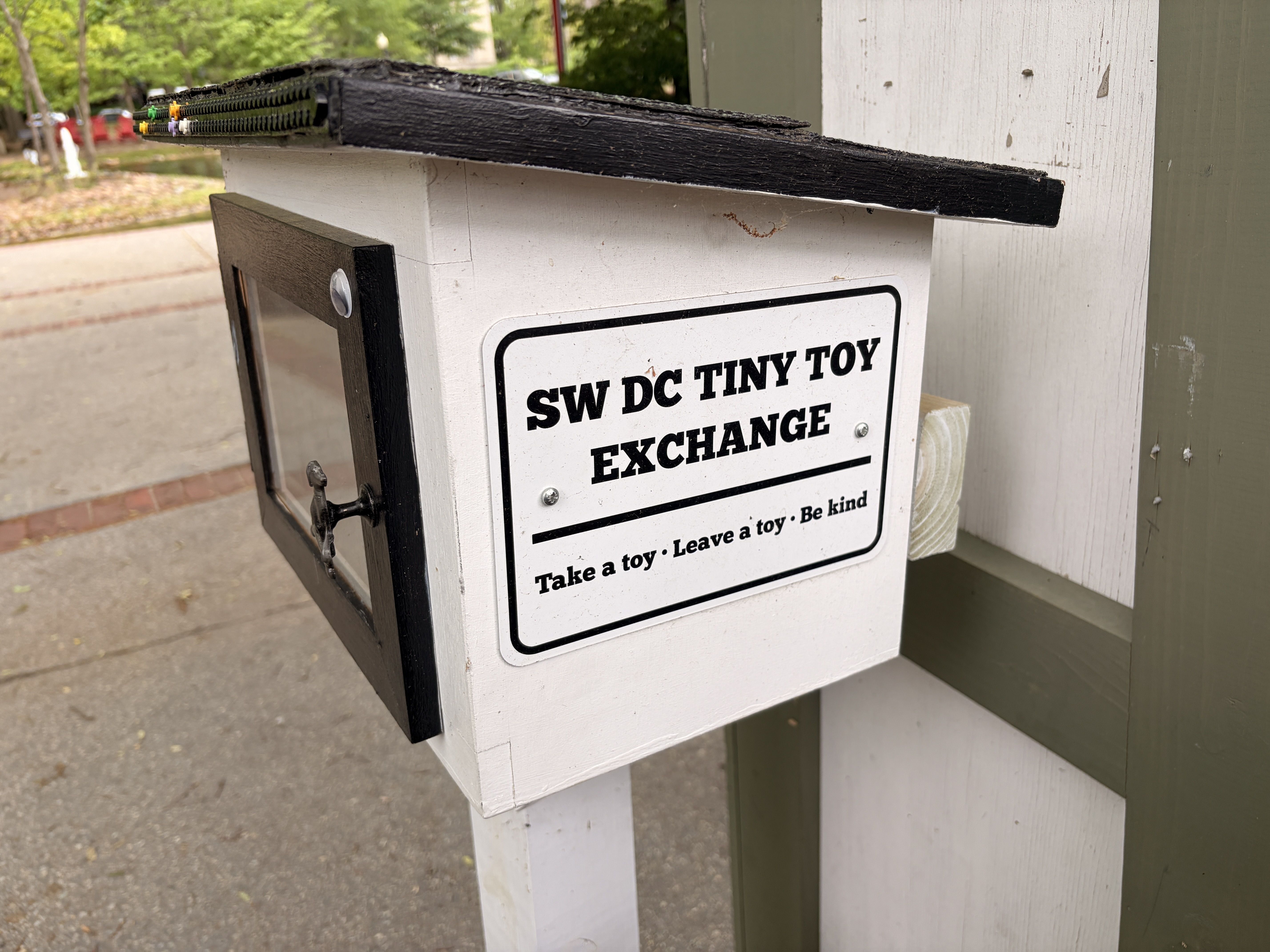 White mailbox mounted on a white post with a black shelf on top; a long black board extends to the right, ending in a row of colorful push pins. The sign reads "SW DC TINY TOY EXCHANGE".