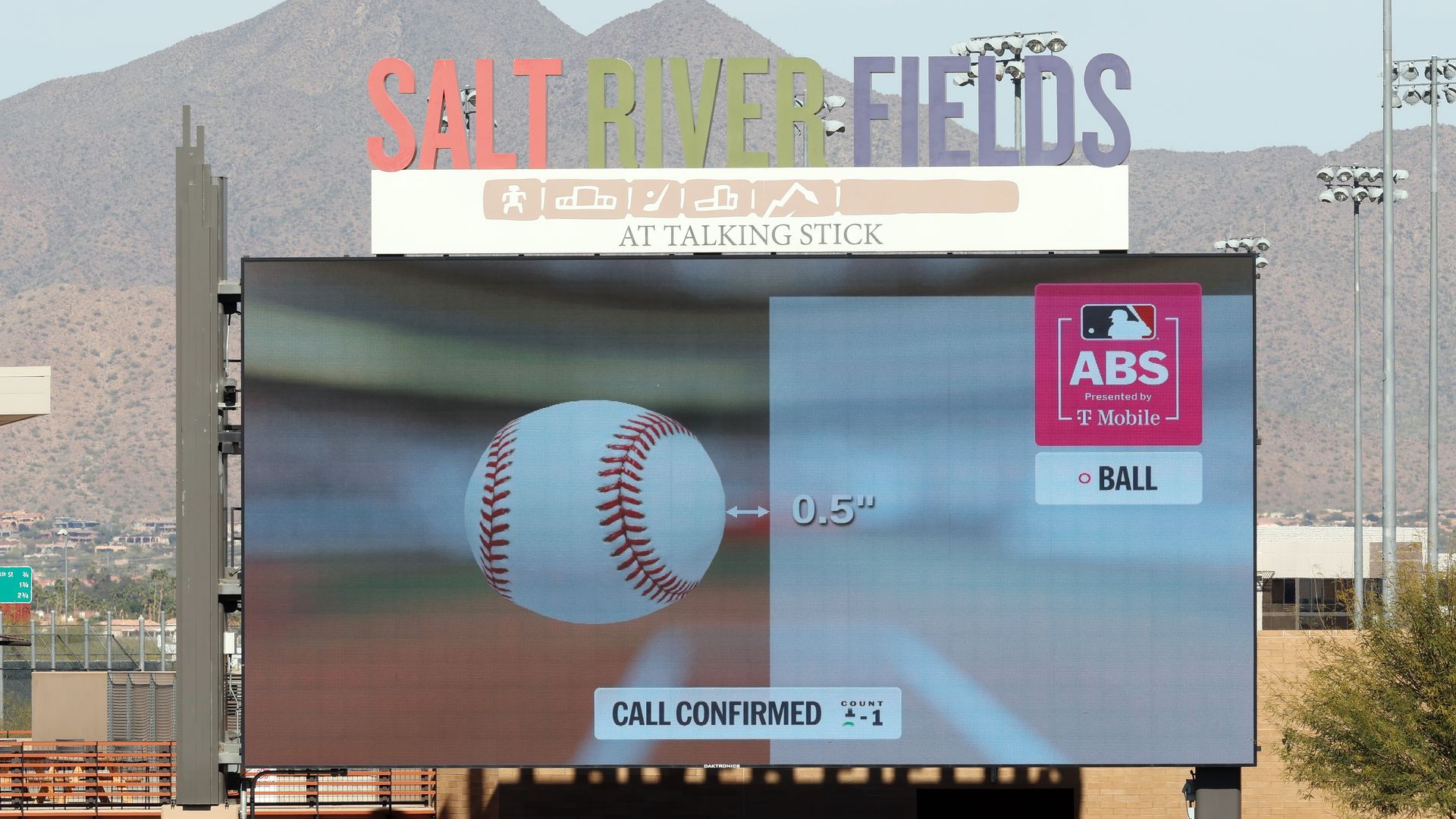 Wide view of Salt River Fields with a tall multicolor sign; a giant video board shows a baseball, 0.5" marker, ABS by T-Mobile logo, mountains in the background, and a crowd below.
