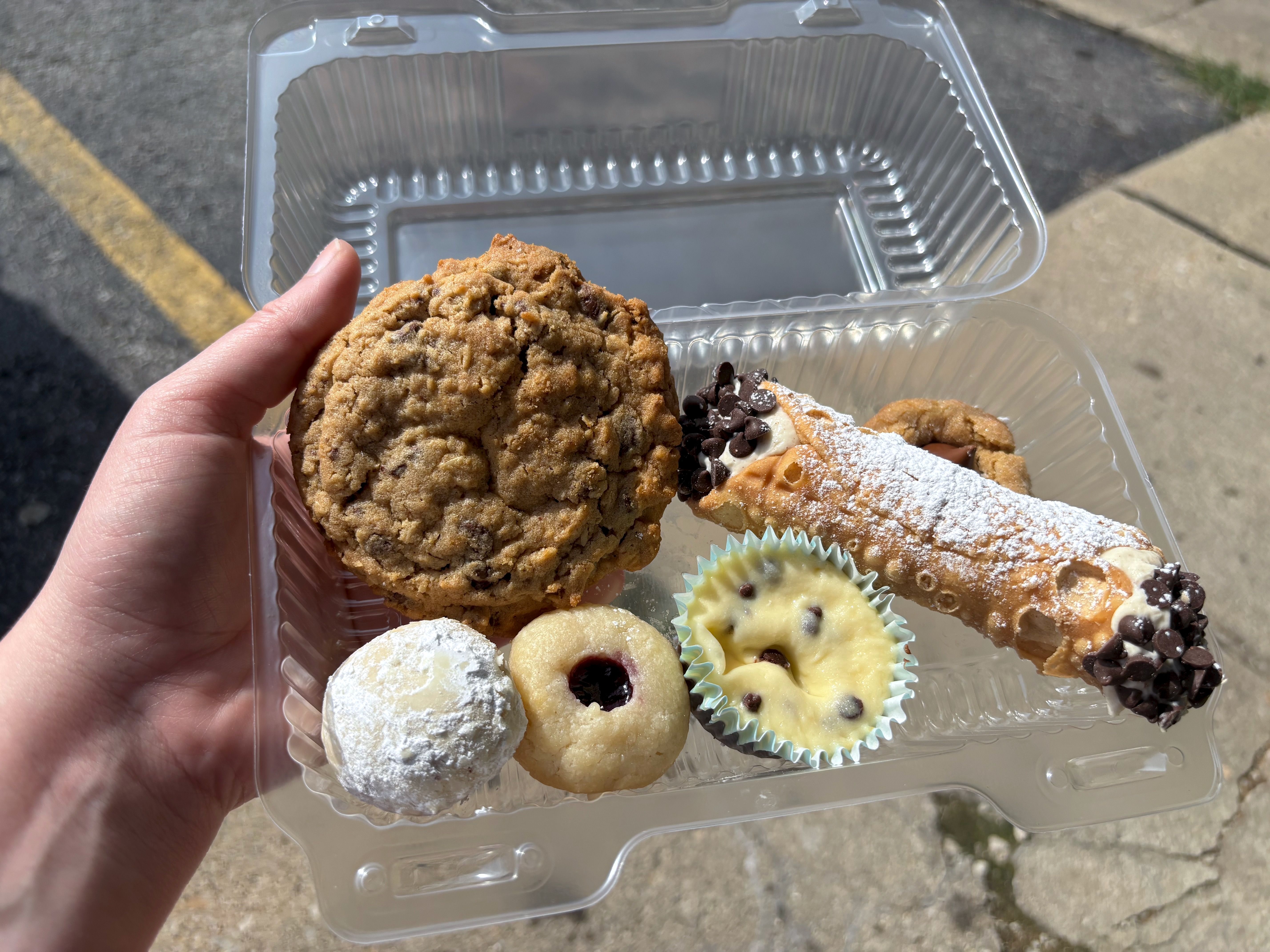 Clear plastic container held in hand with assorted cookies and pastries including chocolate chip, powdered sugar cookie, jam-filled cookie, mini chocolate chip cheesecake, and cannoli dusted with powdered sugar.