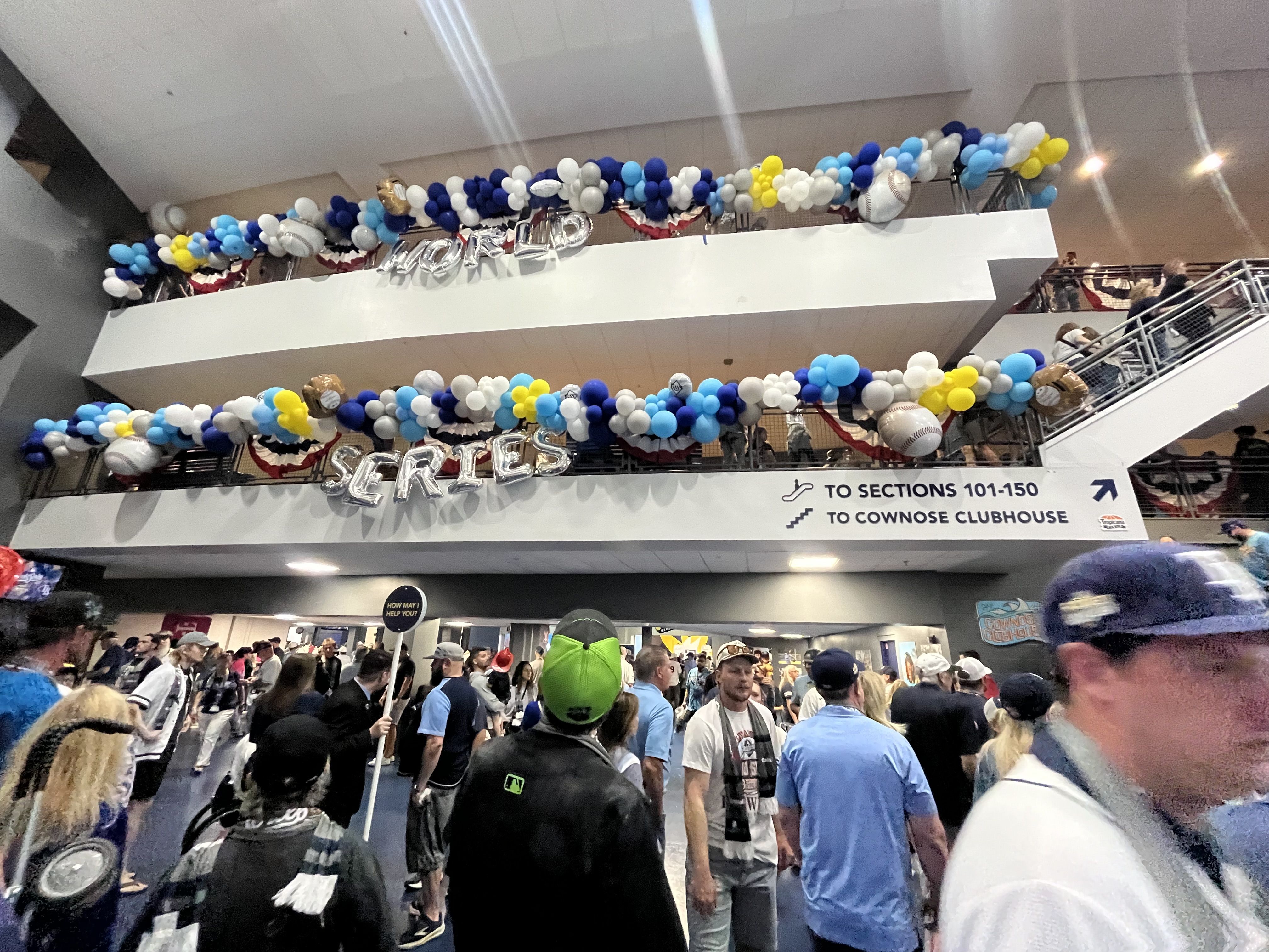 Busy stadium with blue, white, gray, and yellow balloon garlands along railings. Large silver foil letters spell "WORLD" above as crowds fill lower levels and signs direct visitors.