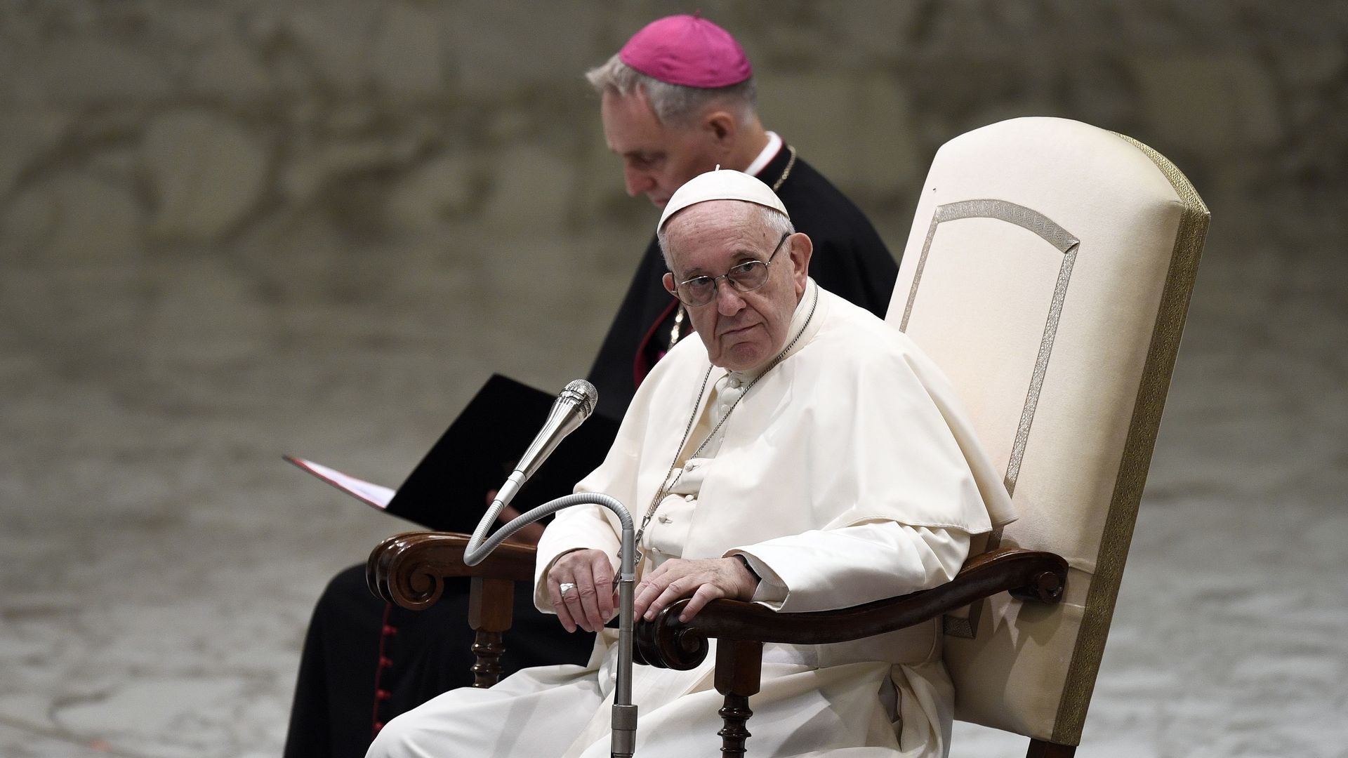The pope sitting in a chair looking to the side, looking serious.