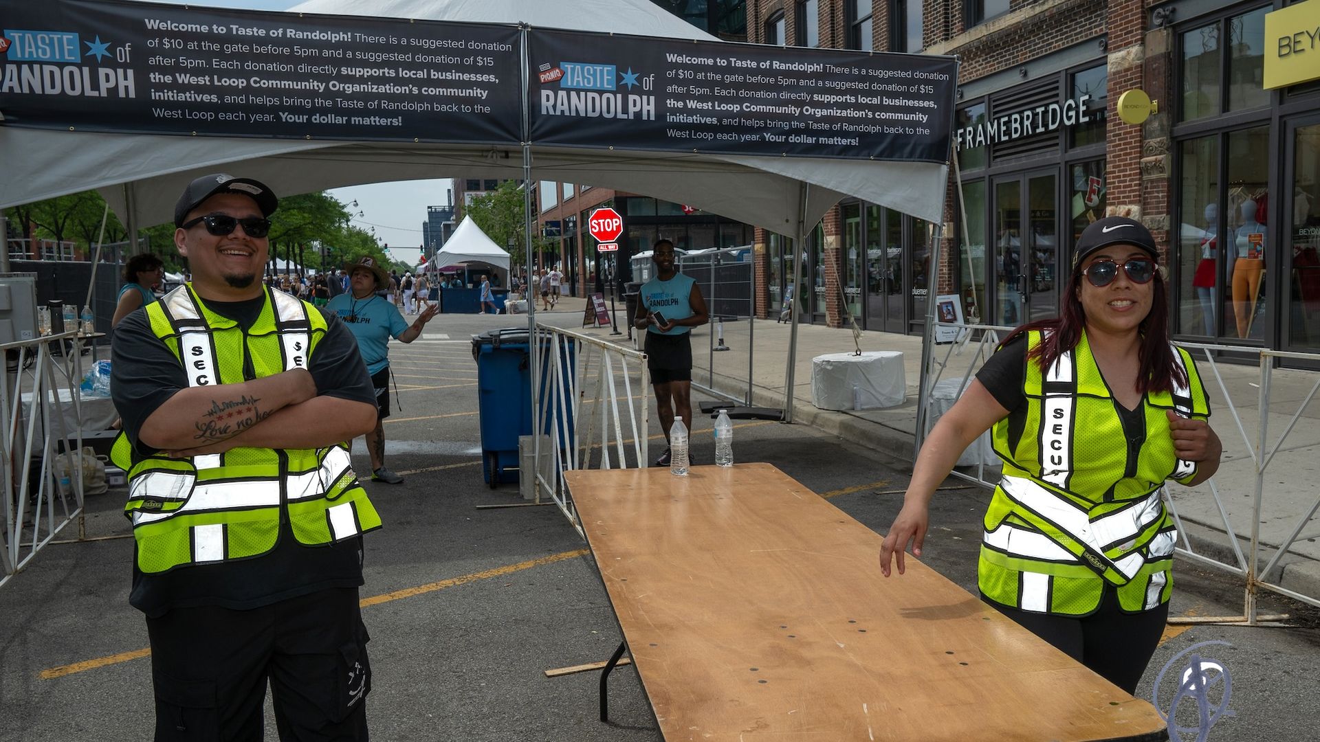 Two smiling security volunteers in neon yellow vests stand at a wooden table under a white tent, with a "Taste of Randolph" banner behind and a Stop sign nearby.