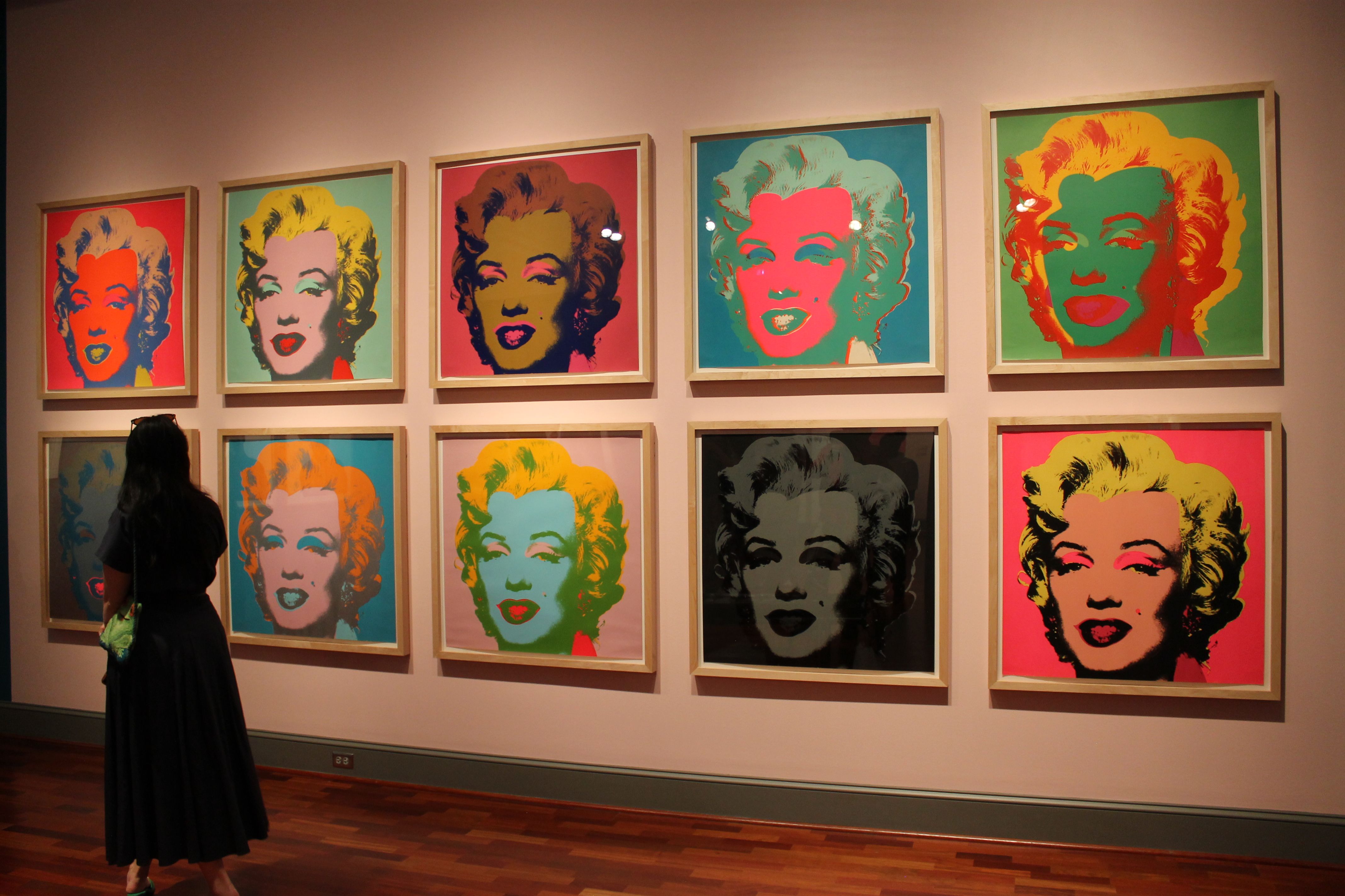 Gallery wall featuring a row of Warhol-style Marilyn Monroe portraits in vivid color blocks; a person in a dark dress stands at left, observing the framed artworks under warm gallery lighting.