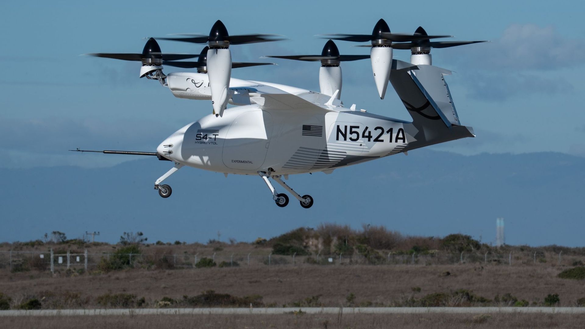 White experimental hybrid VTOL aircraft with registration N5421A hovering over a grassy field, featuring black propellers and an American flag on the side under a clear blue sky.
