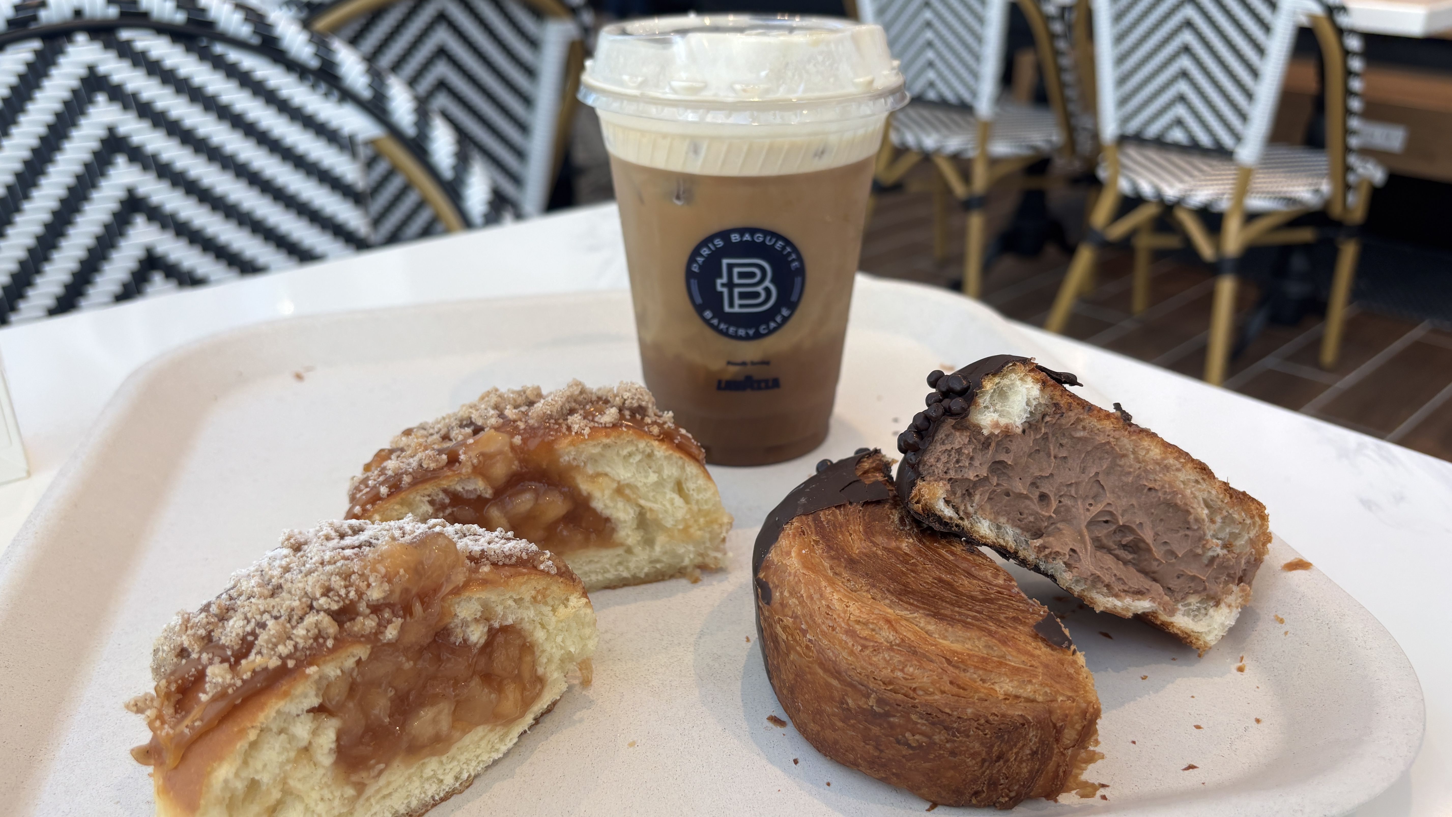 A to-go iced coffee with a lid sits on a white tray beside two pastries (apple caramel danishes) in a cafe with black-and-white chevron-pattern chairs in the background.