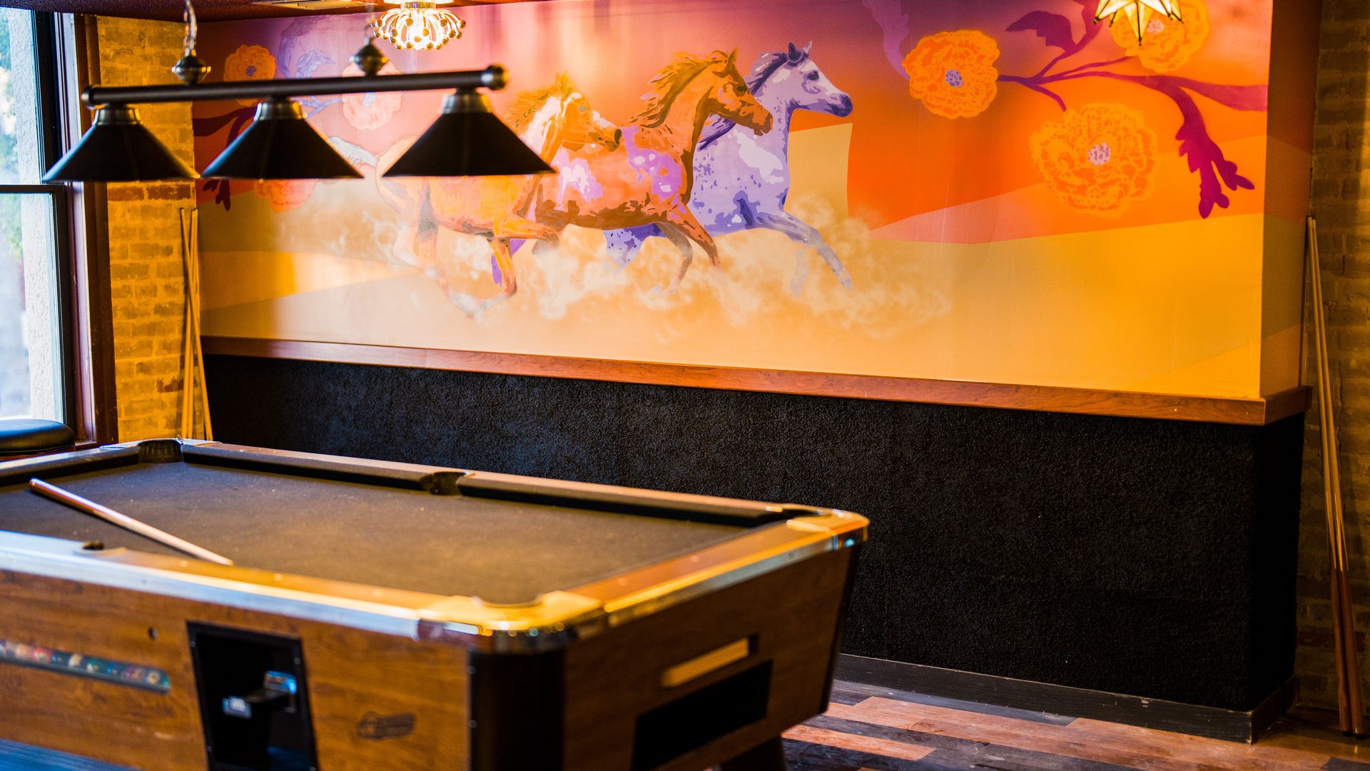 A pool table is near a wall with a mural of horses running.
