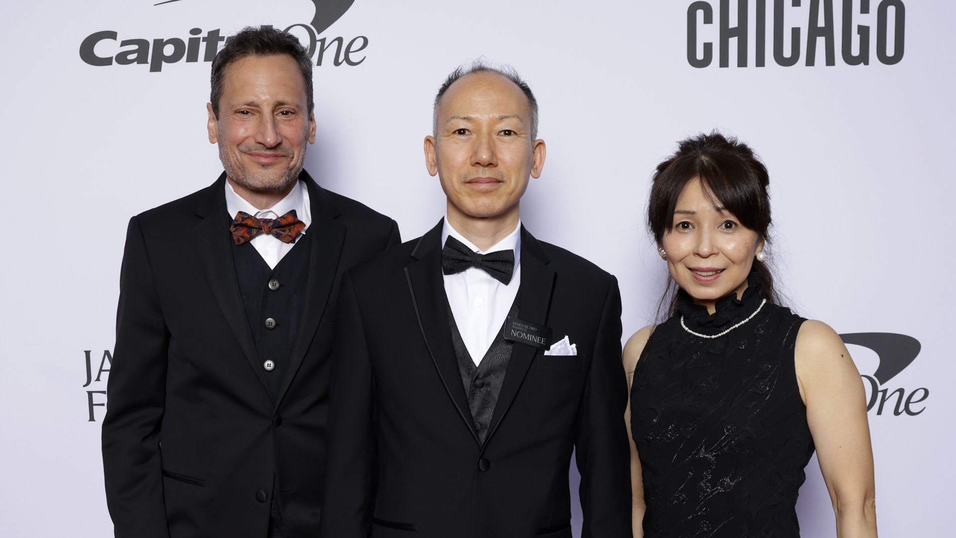 Three people pose on a red carpet in formal wear: two men in black tuxedos with bow ties and a woman in a black dress with a beige clutch; backdrop shows Capital One and CHICAGO logos.